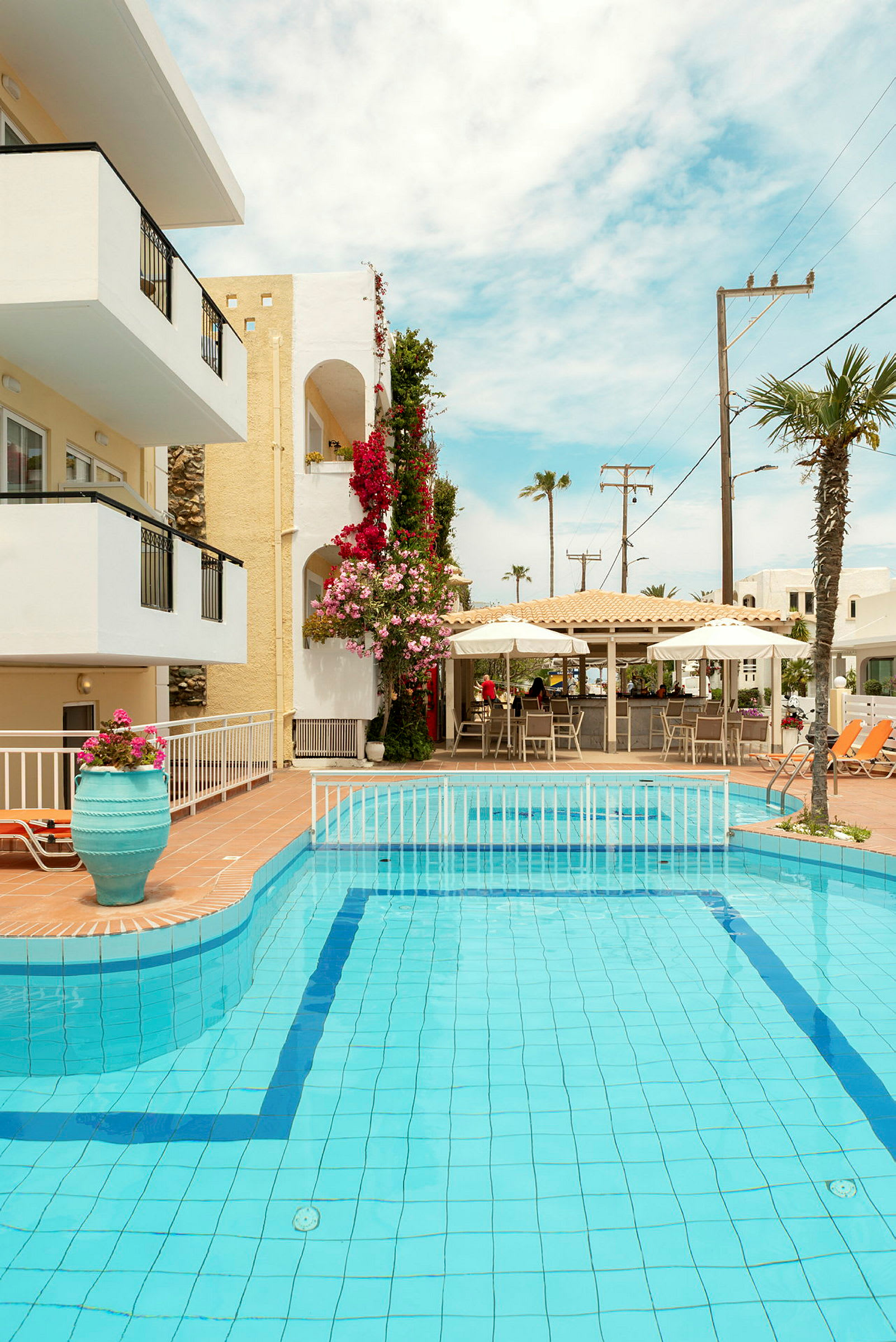 En pool omgivet af terrasseområder, liggestole og bygninger med altaner. Planter og parasoller ses ved poolen. (AI-genereret tekst)