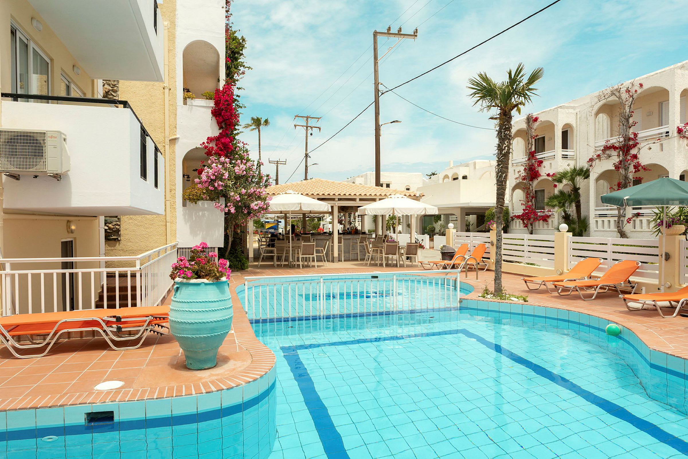 Udendørs swimmingpool med liggestole omgivet af blomsterdekorerede bygninger. (AI-genereret tekst)