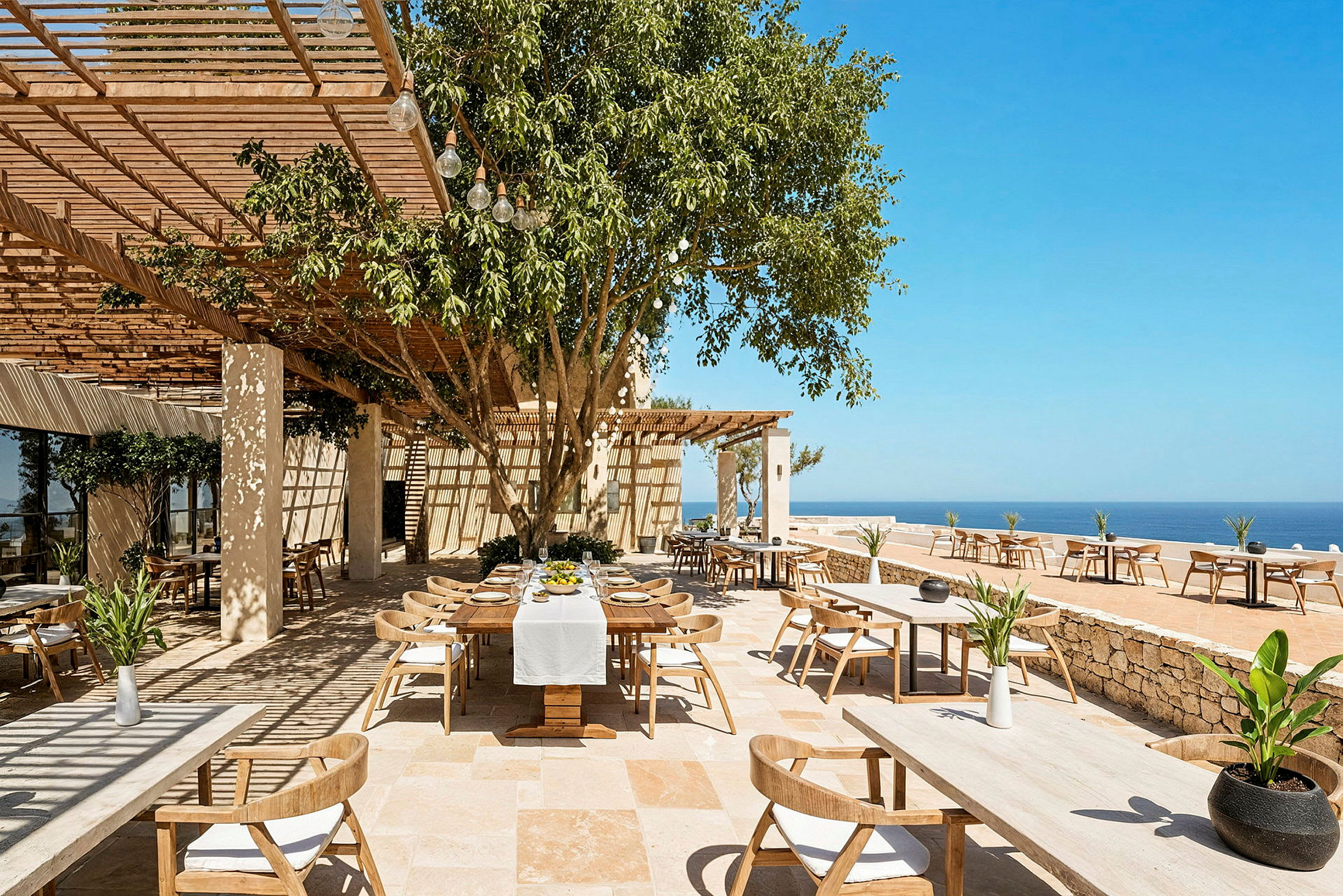 En udendørs restaurant med træmøbler og hvide duge, omgivet af frodig beplantning og med udsigt over havet under en klar blå himmel. (AI-genereret tekst)