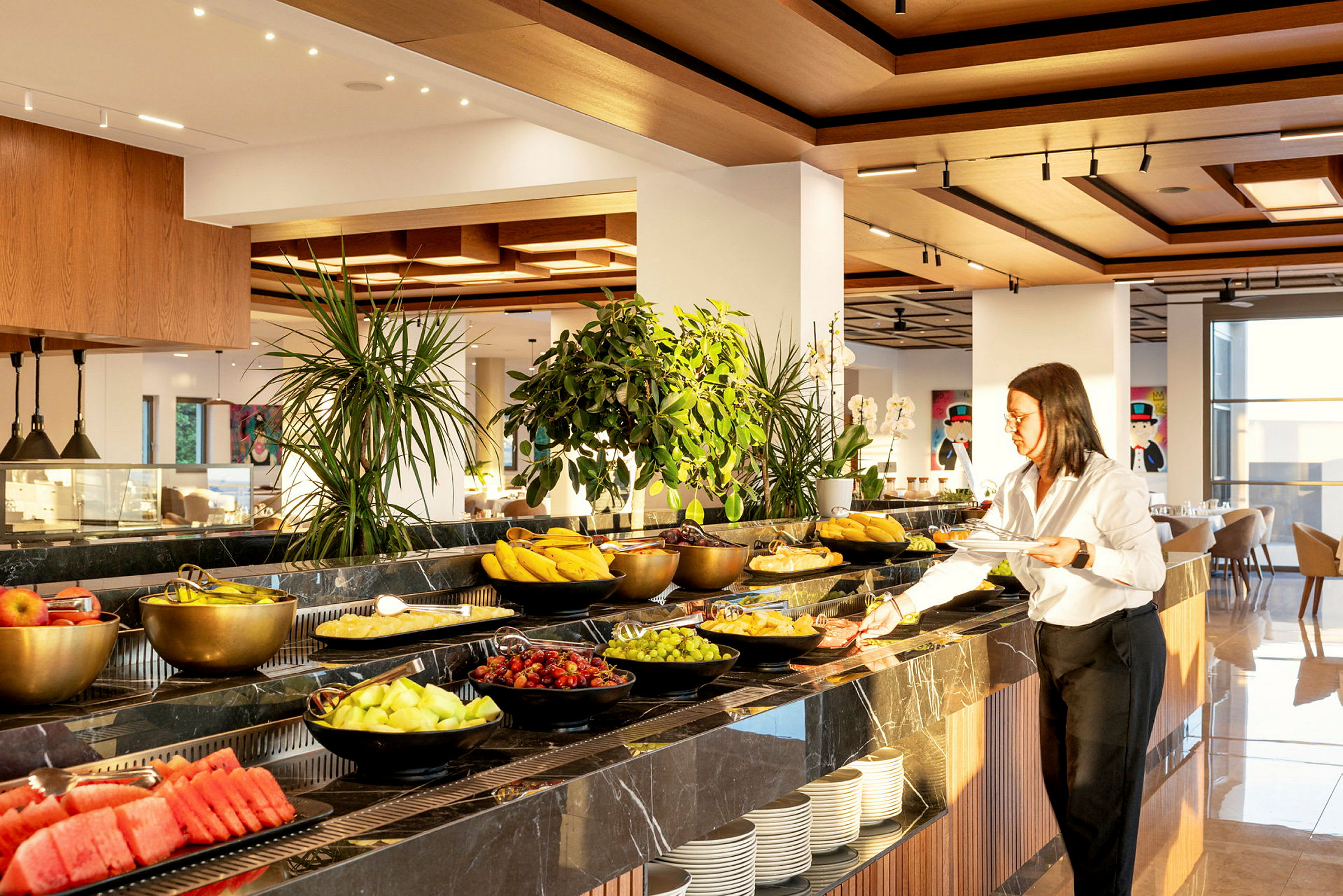En morgenmadsbuffet med forskellige valg af frugter og grøntsager pænt arrangeret i skåle på et bord. En person står ved siden af og tager mad. (AI-genereret tekst)
