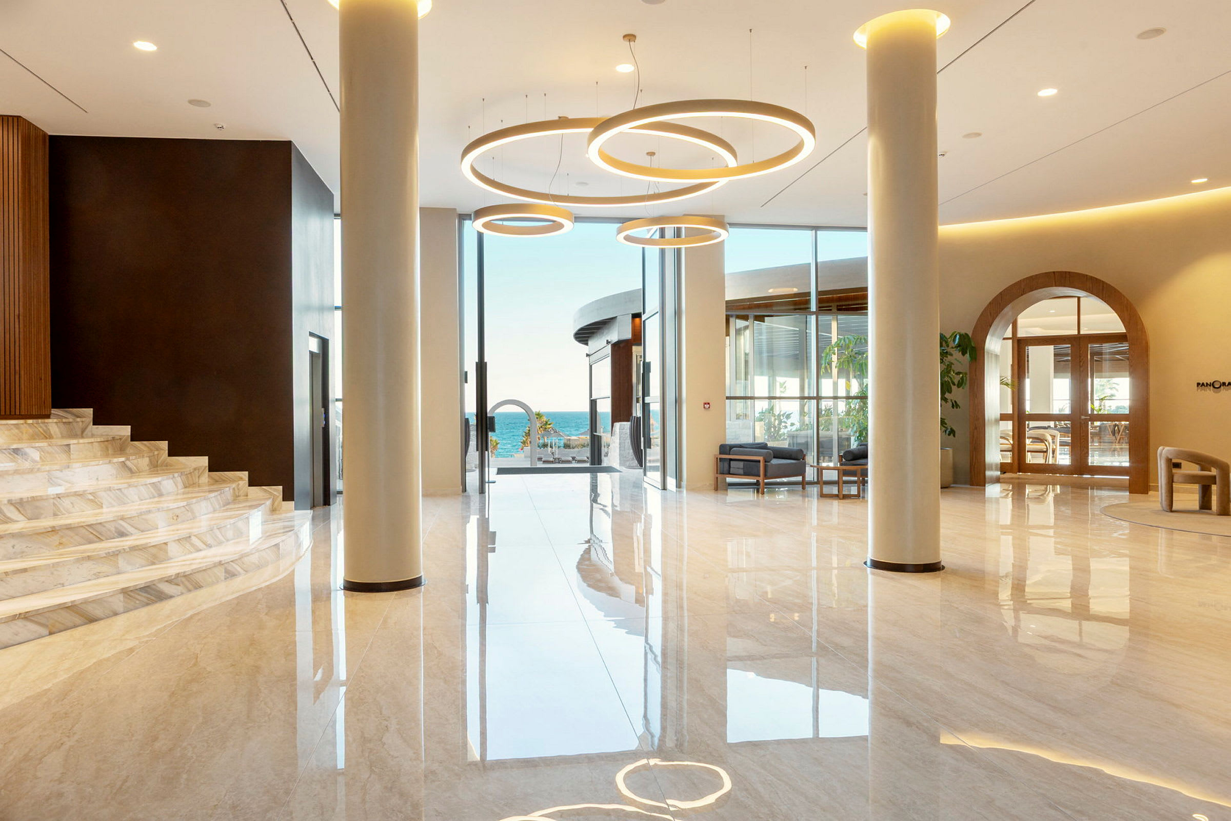 Lys og moderne lobby med store glaspartier og udsigt til havet samt elegante lamper i loftet. (AI-genereret tekst)