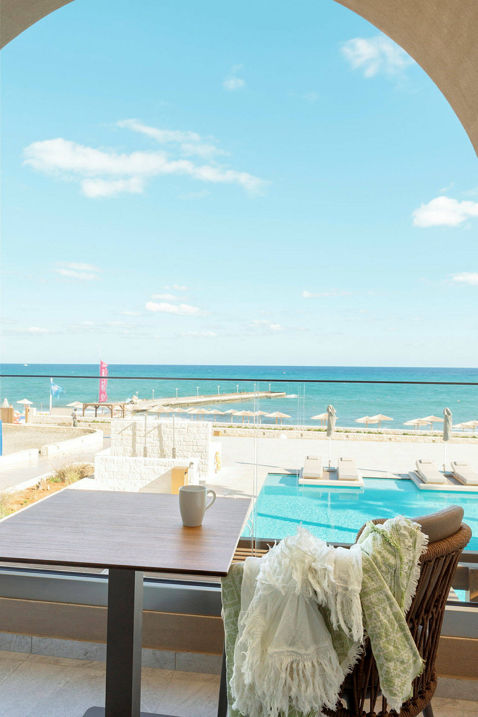 Udsigt over en pool og havet fra en terrasse, med et bord og en stol i forgrunden. (AI-genereret tekst)