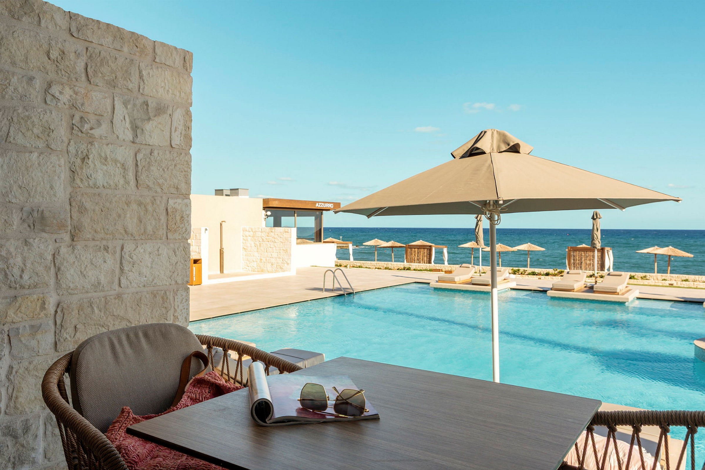 Udsigt fra en terrasse mod en pool med parasoller og liggestole, med havet i baggrunden. (AI-genereret tekst)