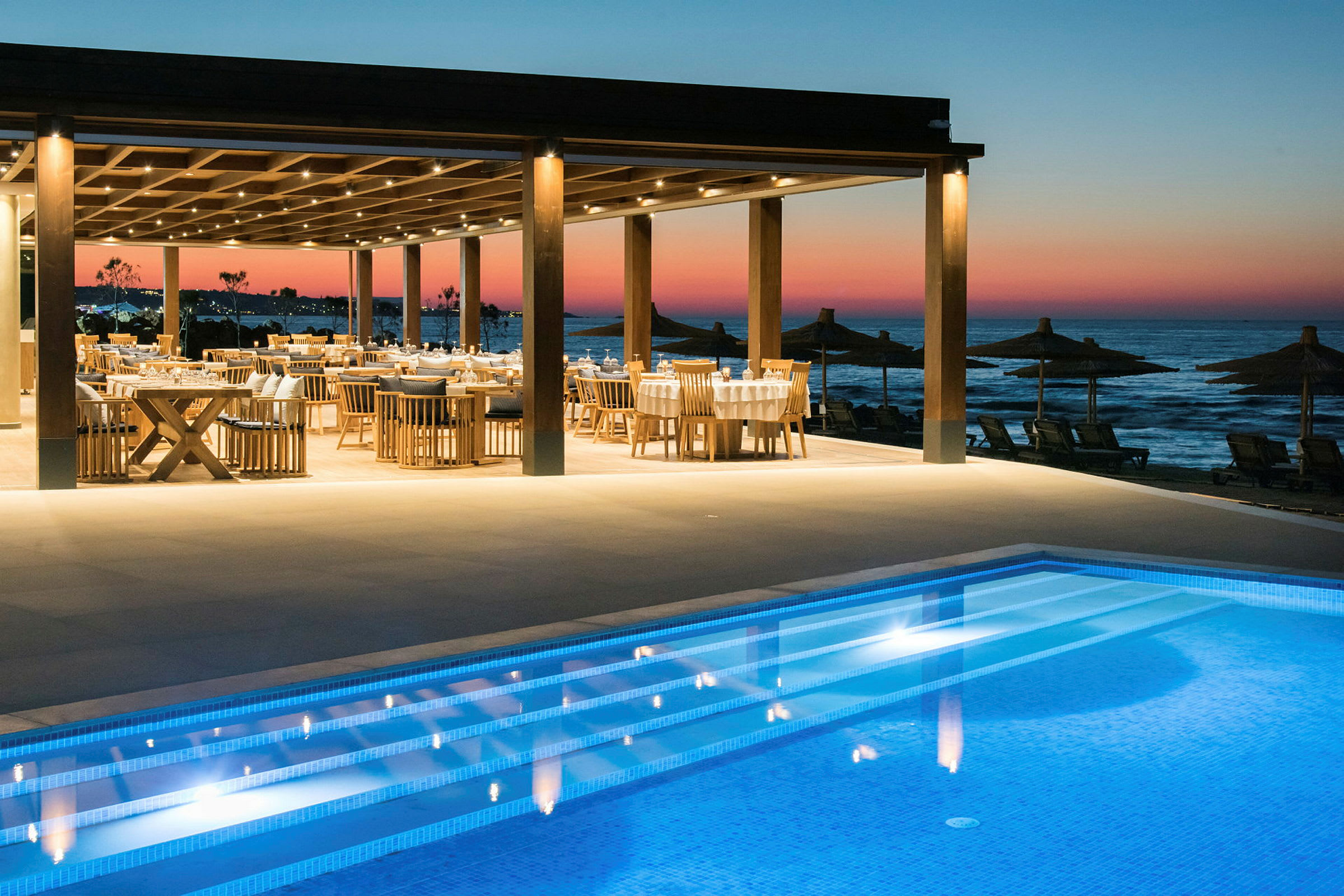 Restaurant med borde og stole nær en pool og havet ved solnedgang. (AI-genereret tekst)