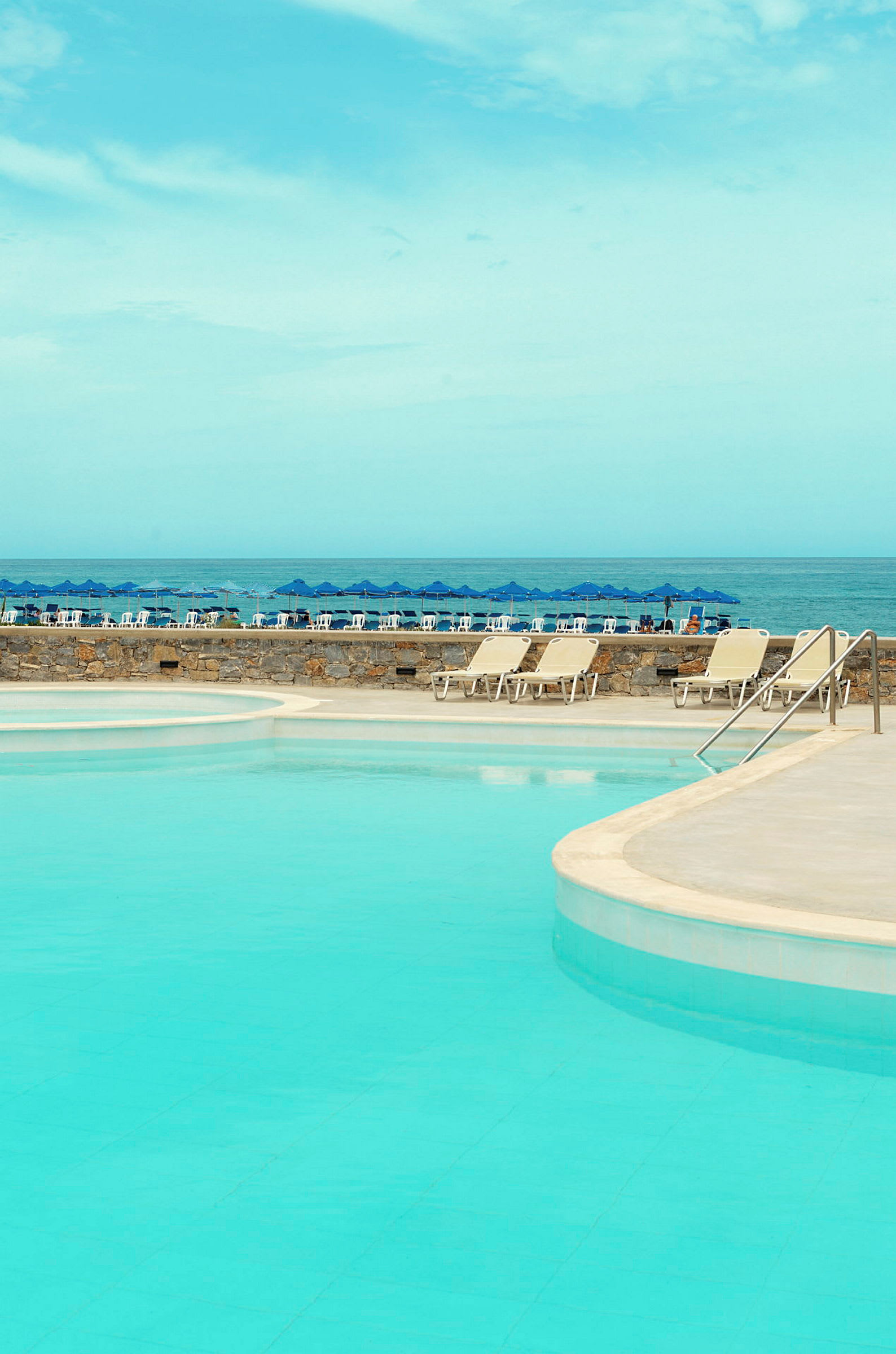 En pool med klart blåt vand og liggestole ved kanten, samt en blå himmel og blå parasoller ved havet i baggrunden. (AI-genereret tekst)