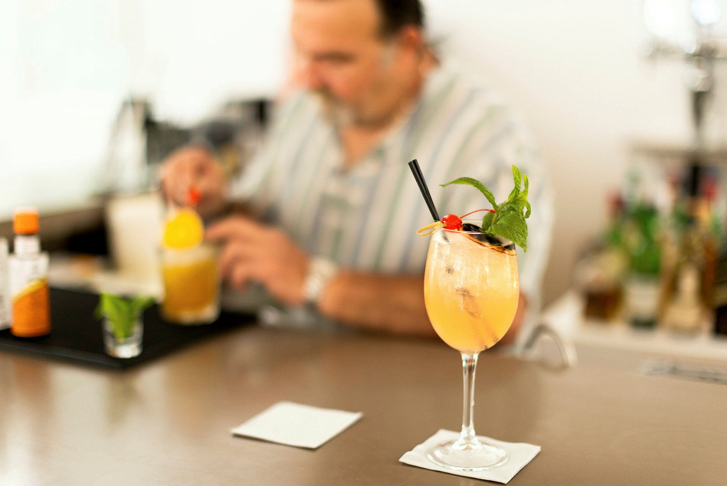 Et cocktailglas med en farverig drik, toppet med is, mynte og et kirsebær, står på en bardisk. En person i baggrunden blander en drink. (AI-genereret tekst)