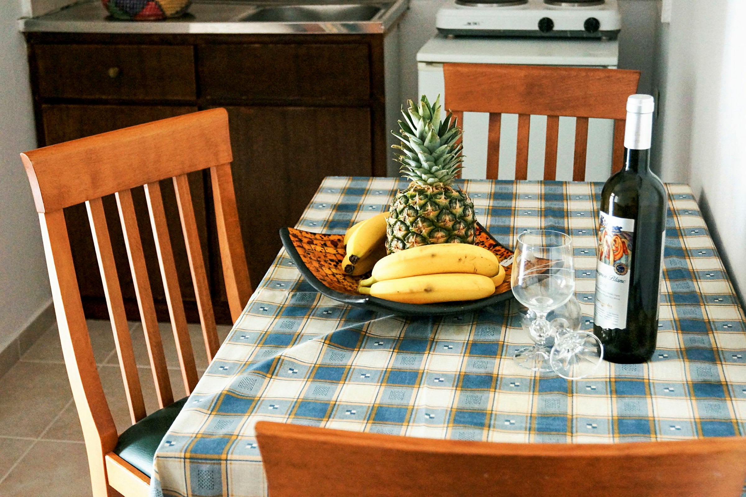 Et spisebord med en frugtskål med ananas og bananer, samt en flaske vin og to vinglas. (AI-genereret tekst)