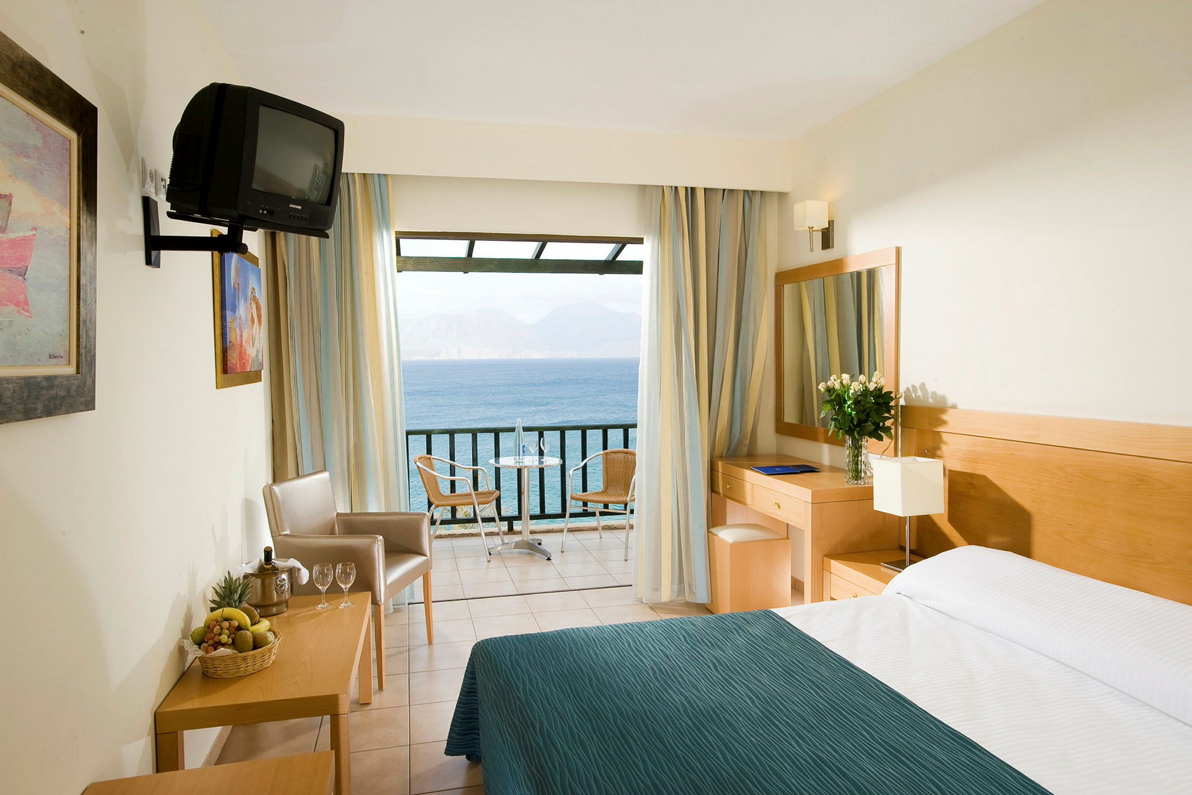 Et hotelværelse med en seng, et skrivebord, stole og en balkon, der tilbyder udsigt over havet. (AI-genereret tekst)