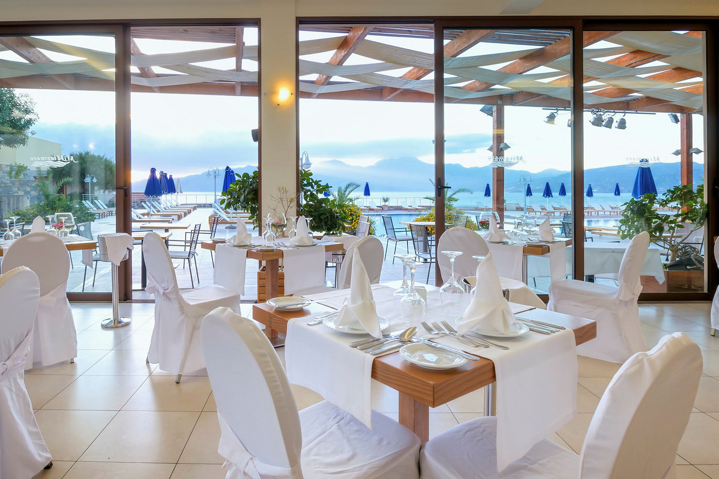 En restaurant med dækkede borde og stole, ved en terrasse med udsigt over bjerge og vand. (AI-genereret tekst)