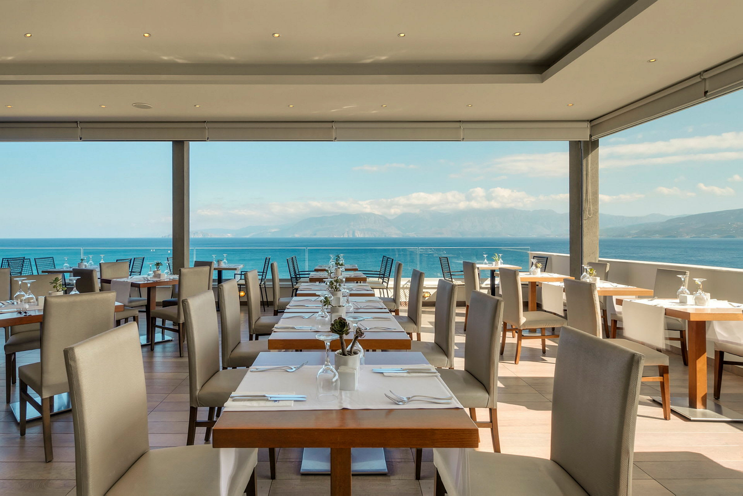 Udsigt over en restaurant med opdækkede borde og stole, med havet og bjergene i baggrunden. (AI-genereret tekst)