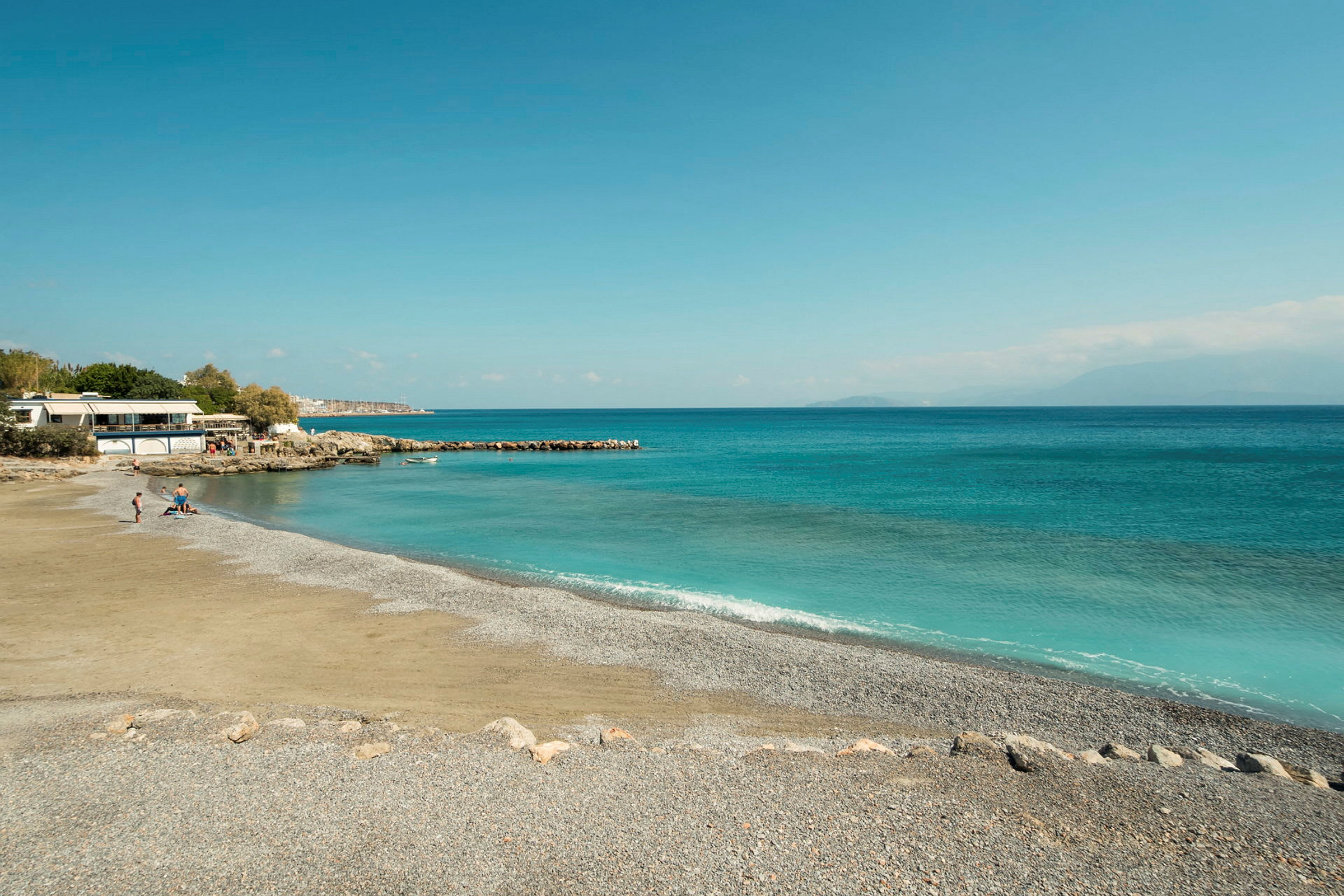 Strand med småsten og klart blå vand, med nogle mennesker nær en bygning. (AI-genereret tekst)