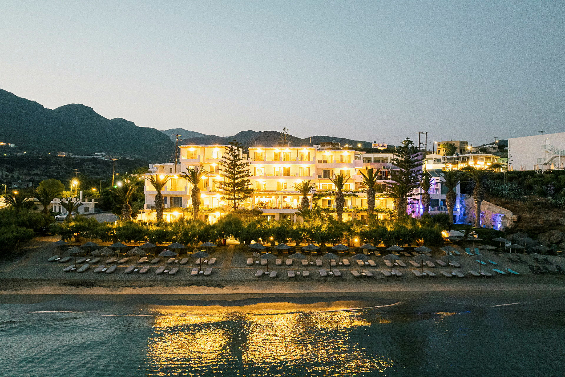Hotel ved havet med oplyst facade, omgivet af palmer og strand med liggestole. (AI-genereret tekst)