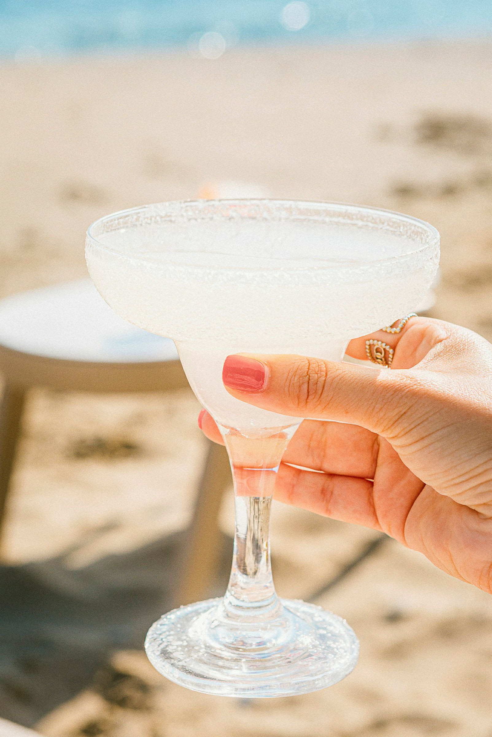 En hånd holder et glas margarita med is ved siden af en strand. (AI-genereret tekst)