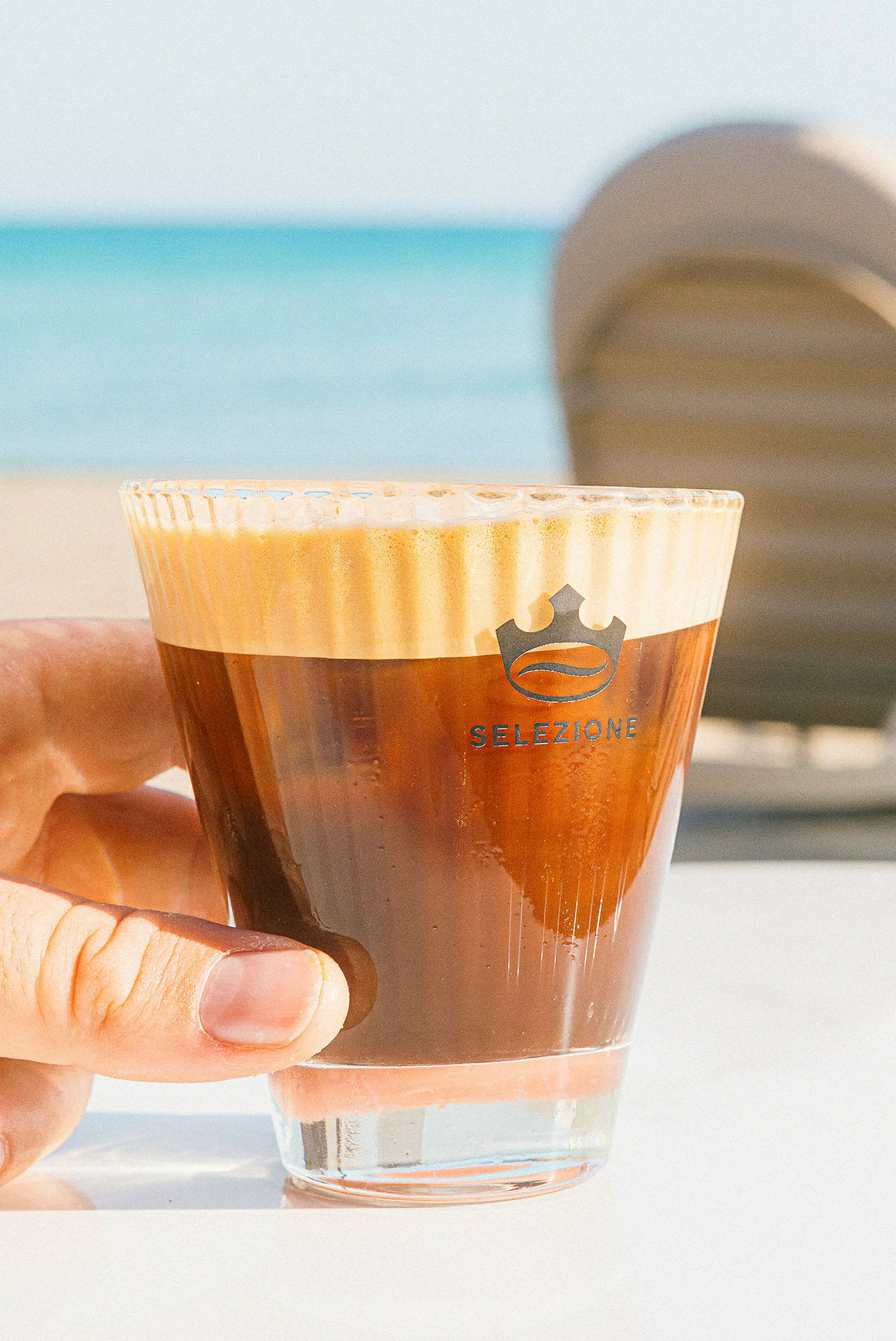 Nærbillede af en hånd, der holder et glas med kaffe, stranden ses i baggrunden. (AI-genereret tekst)