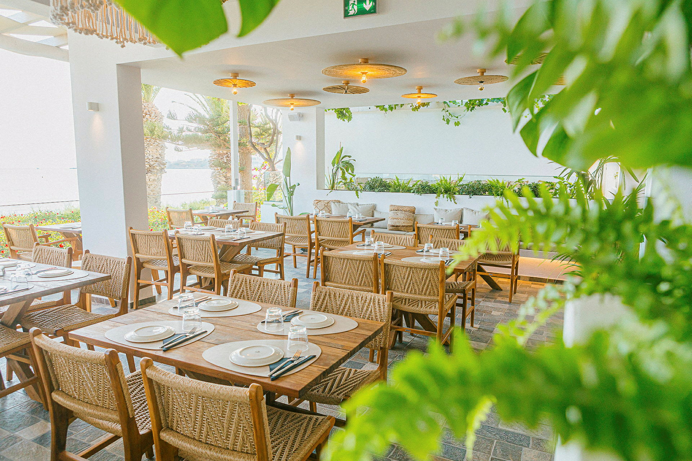 En restaurant med træborde og -stole omgivet af grønne planter og med udsigt over havet. (AI-genereret tekst)
