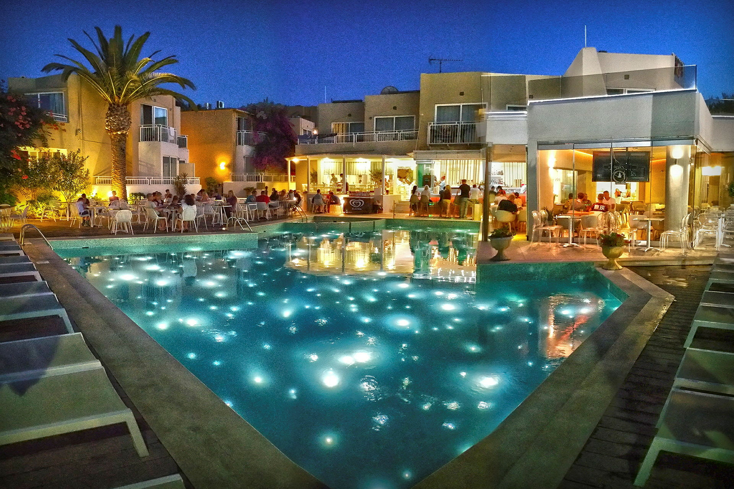 Udsigt over hotelområde med belyst swimmingpool og mennesker ved restaurant om aftenen. (AI-genereret tekst)