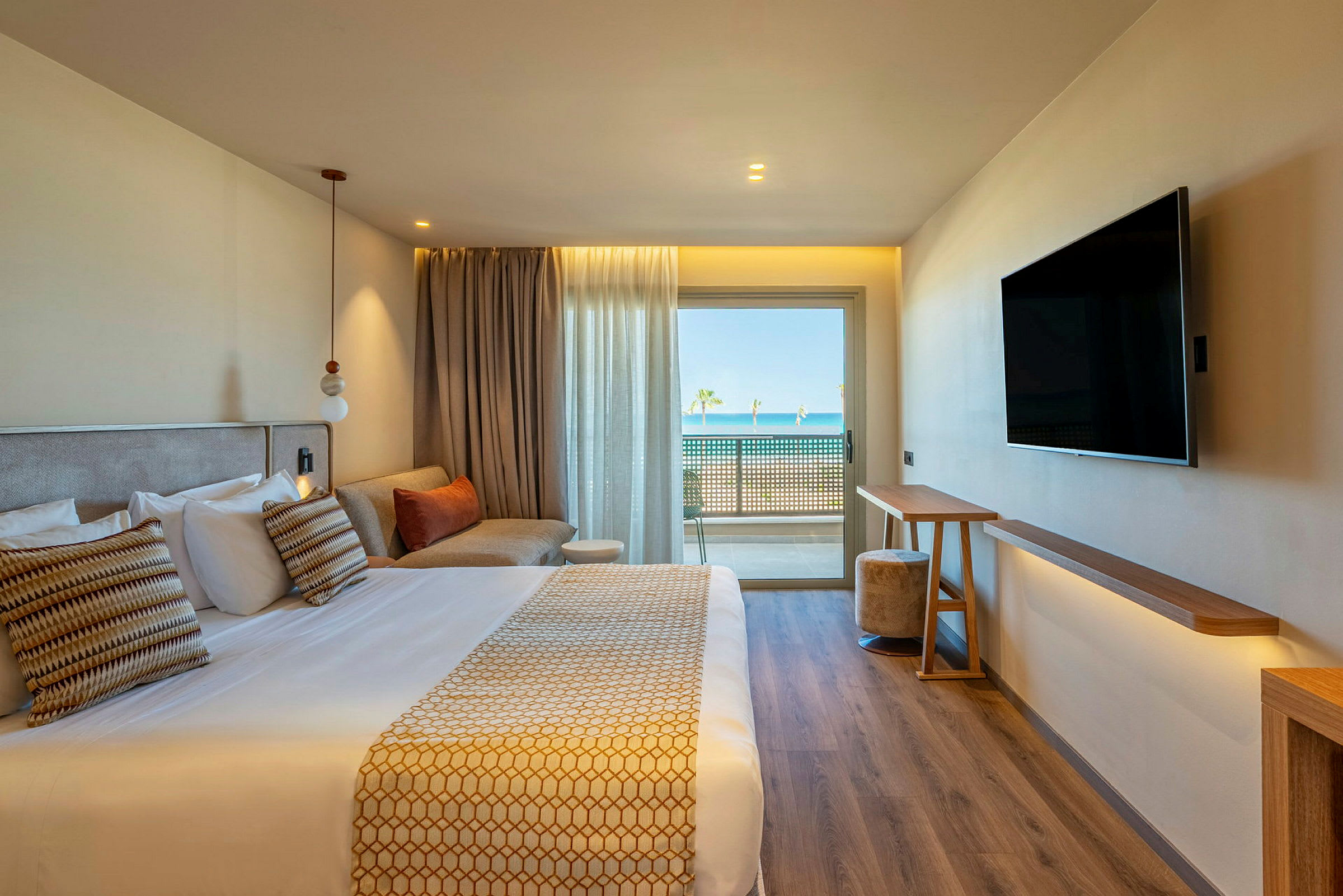Et moderne hotelværelse med en dobbeltseng, en sofa, et vægmonteret TV og store vinduer med udsigt til en balkon og havet i baggrunden. (AI-genereret tekst)