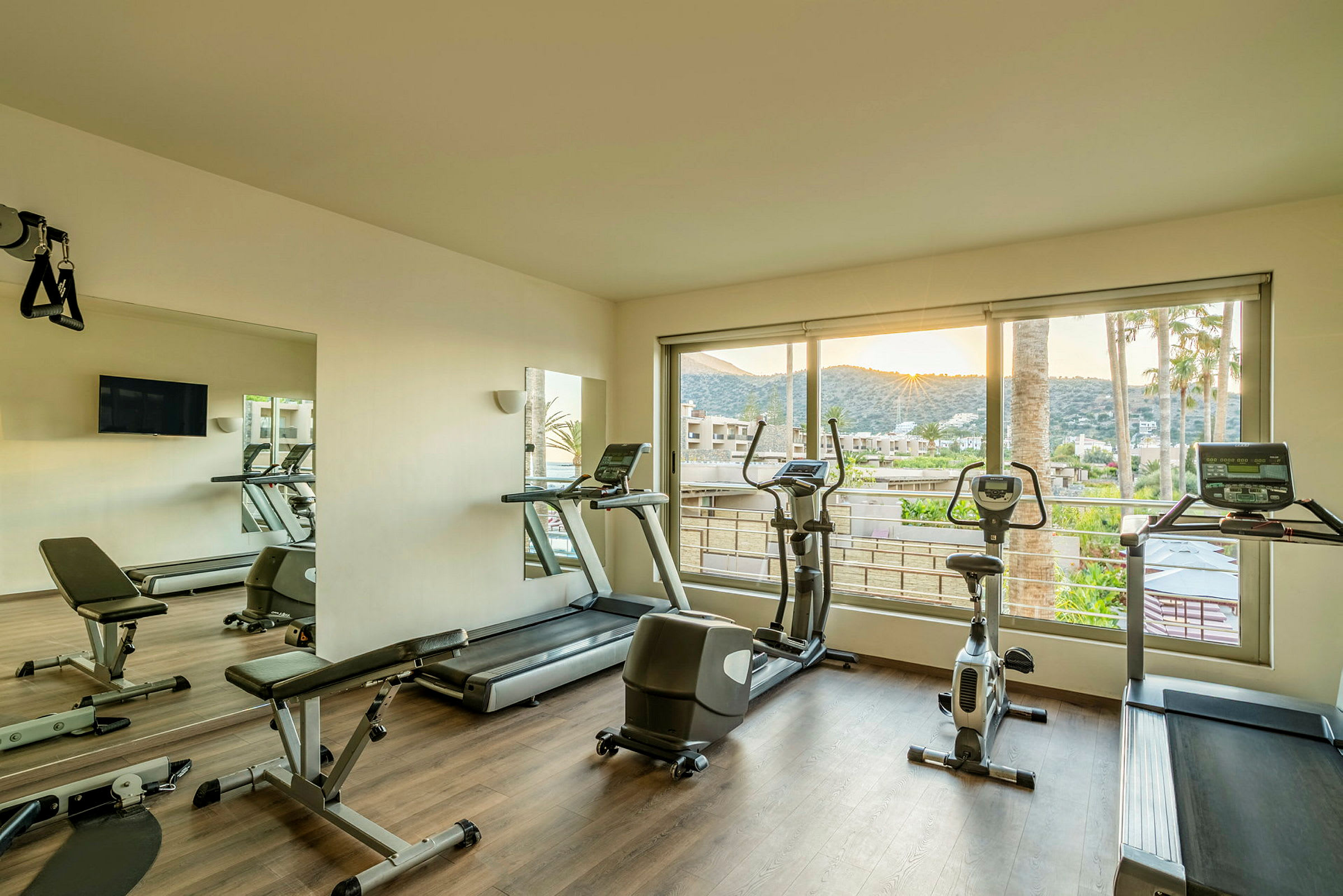 Et træningsrum med fitnessmaskiner som løbebånd og en motionscykel, samt store vinduer med udsigt til solnedgangen og et landskab udenfor. (AI-genereret tekst)