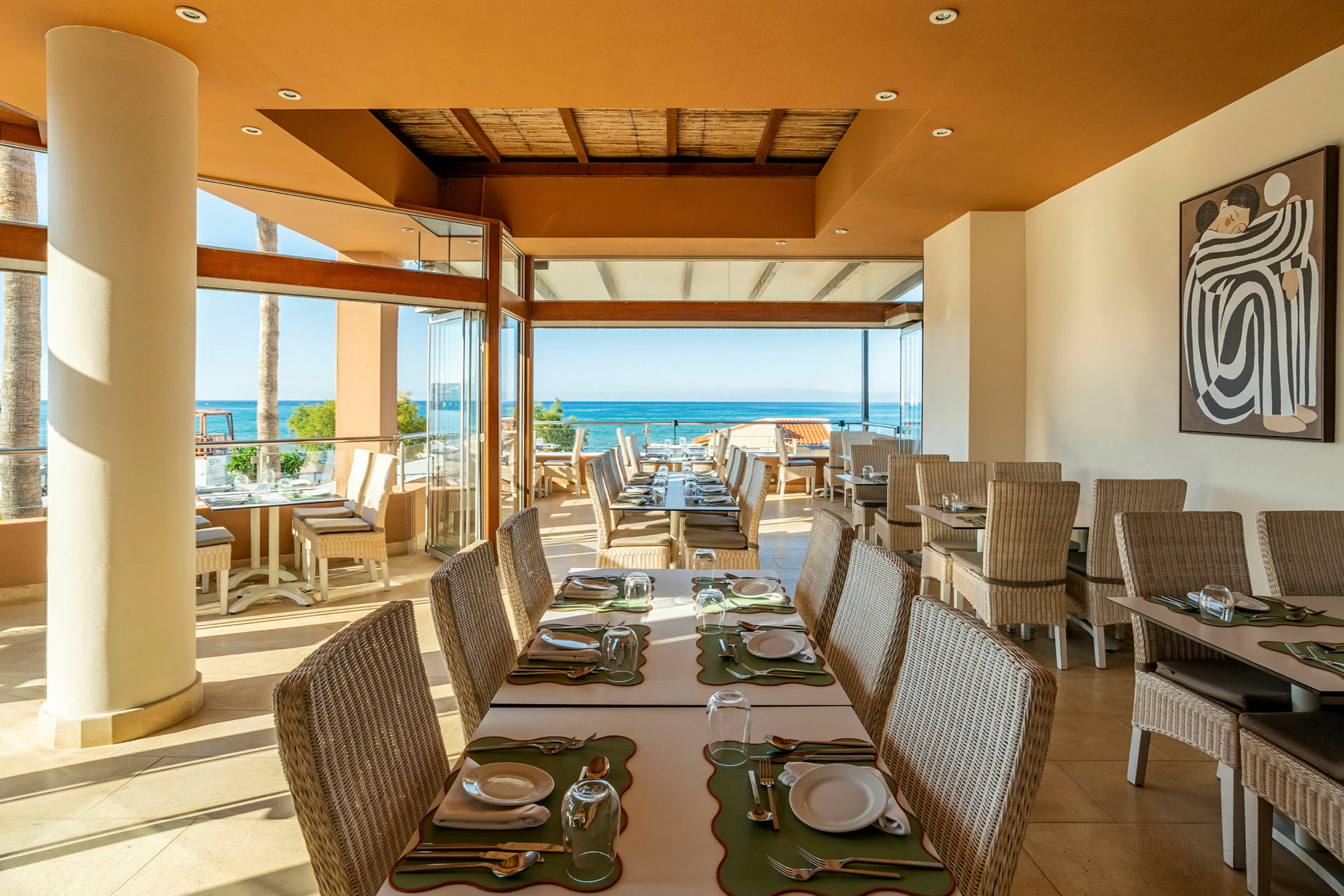 Et solbelyst restaurantområde med borde og stole i rattan, udsigt over havet og et maleri på væggen. (AI-genereret tekst)