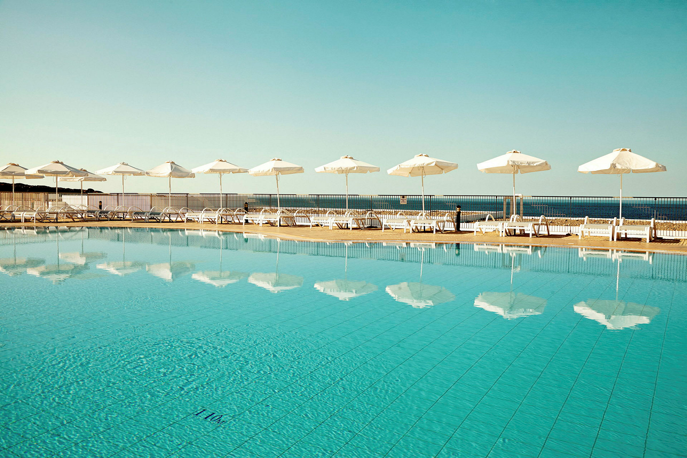 Udendørs pool med klart vand, der reflekterer hvide parasoller, liggestole og havet i baggrunden. (AI-genereret tekst)