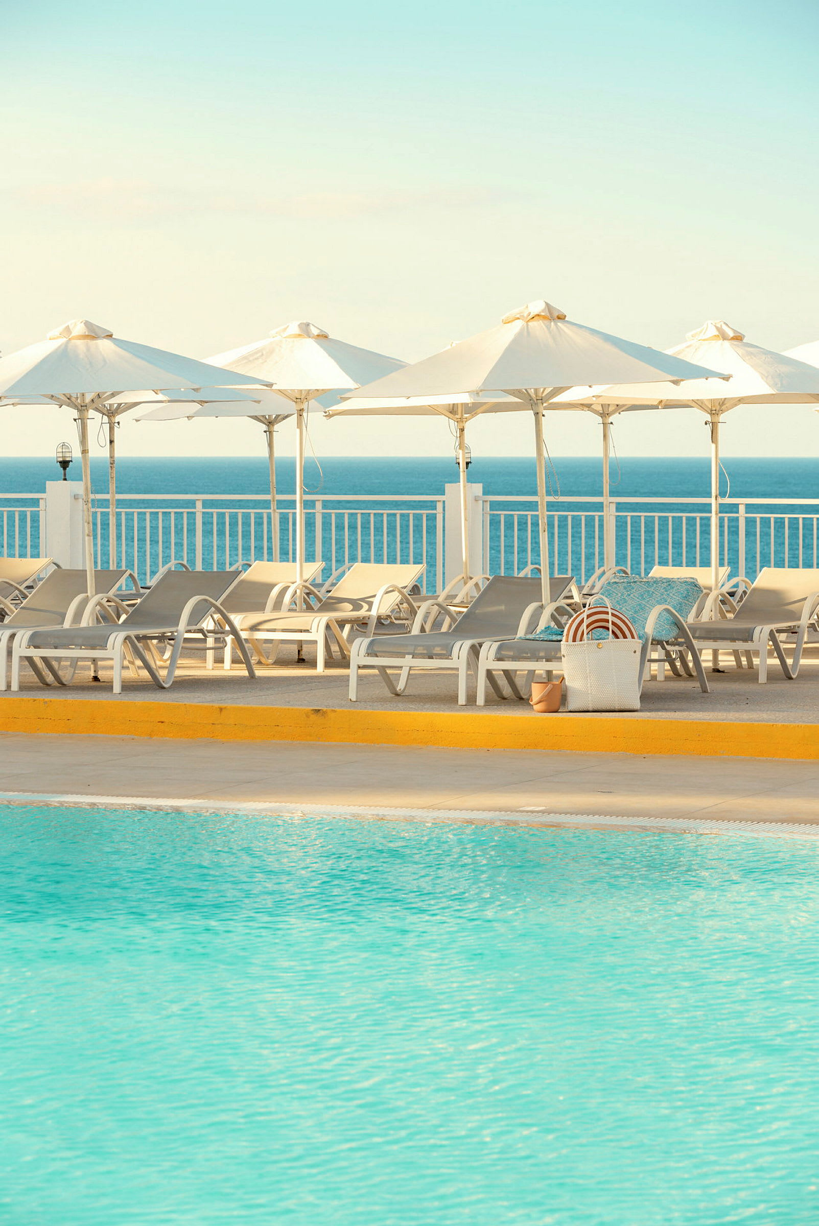 Liggestole og parasoller ved en swimmingpool, med havet i baggrunden. (AI-genereret tekst)