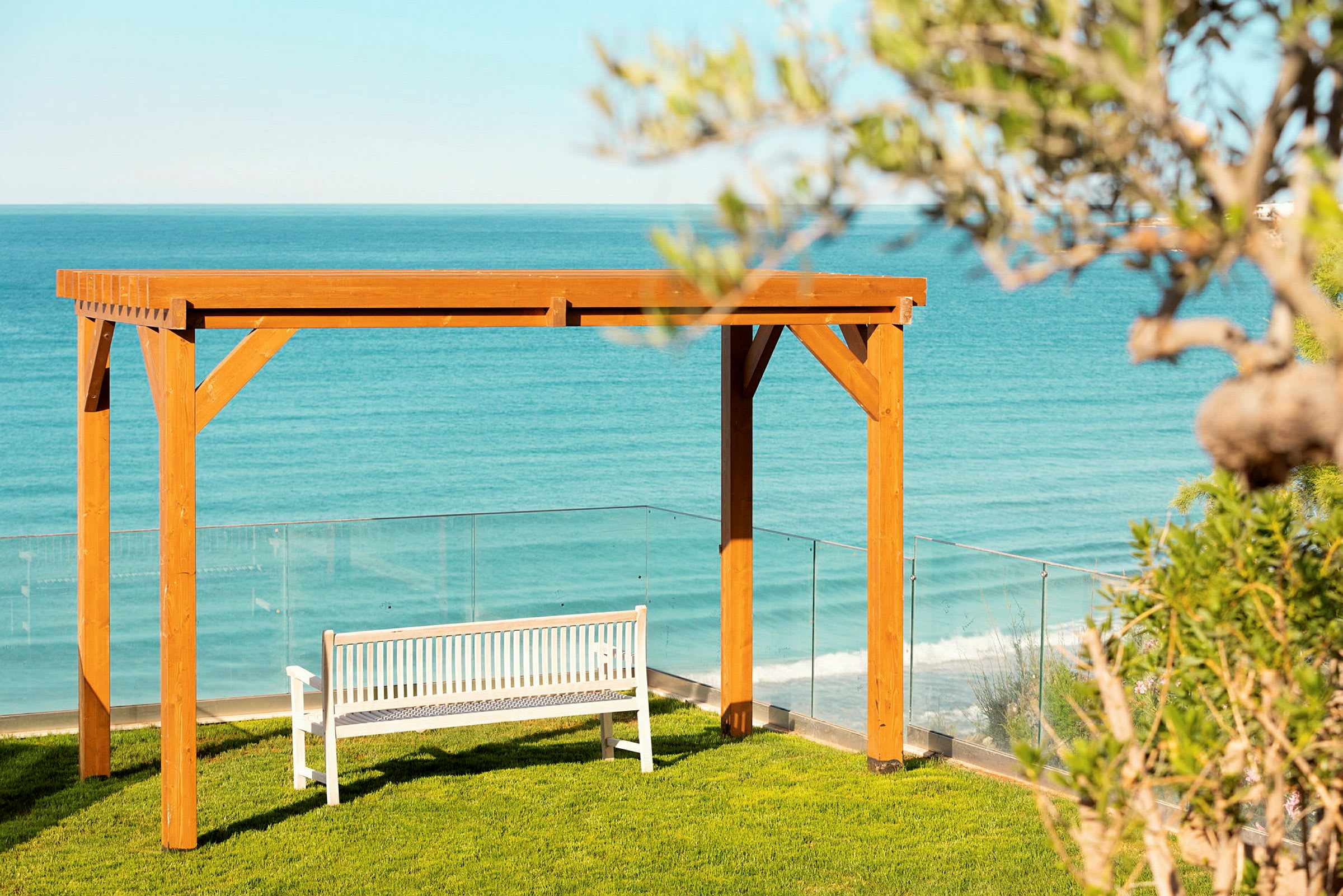 En hvid bænk under en træpergola på en græsplæne med havet i baggrunden. (AI-genereret tekst)