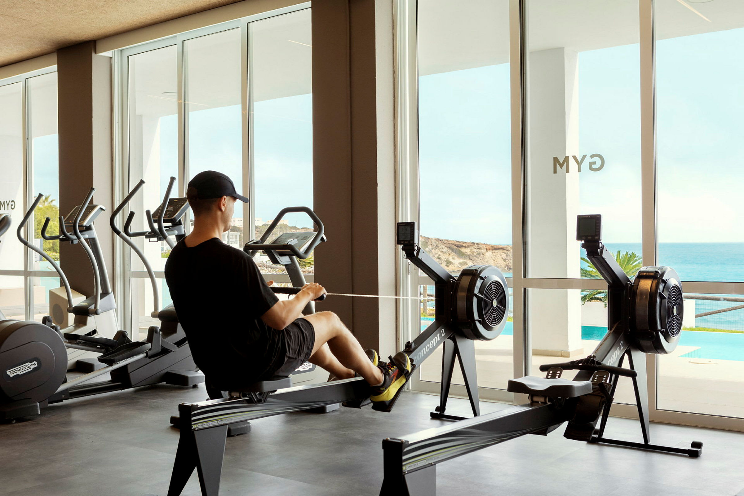 Person træner på en romaskine i et fitnesscenter med havudsigt. (AI-genereret tekst)