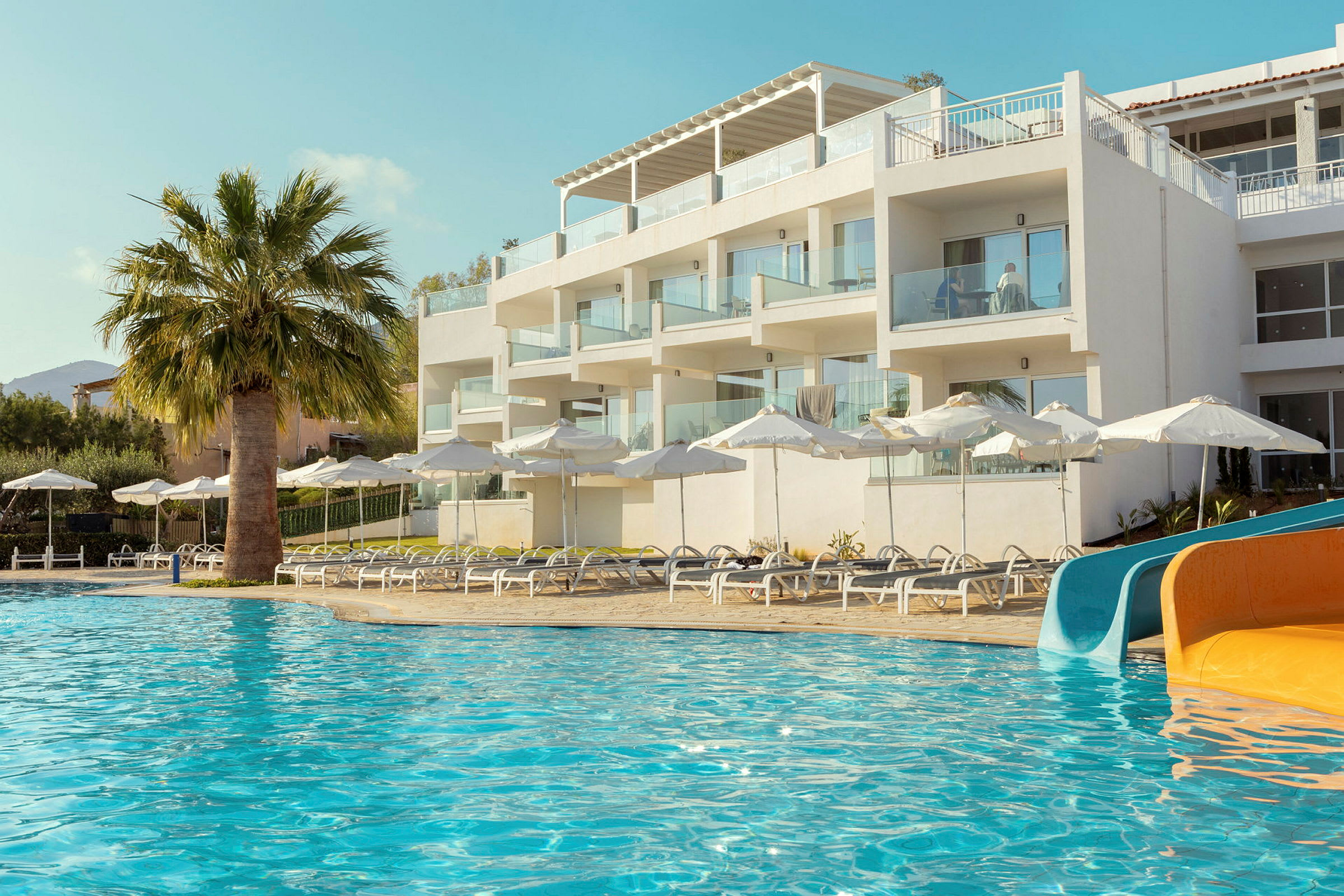 Et hotel med hvide bygninger ved en pool med liggestole, parasoller og vandrutsjebaner. (AI-genereret tekst)