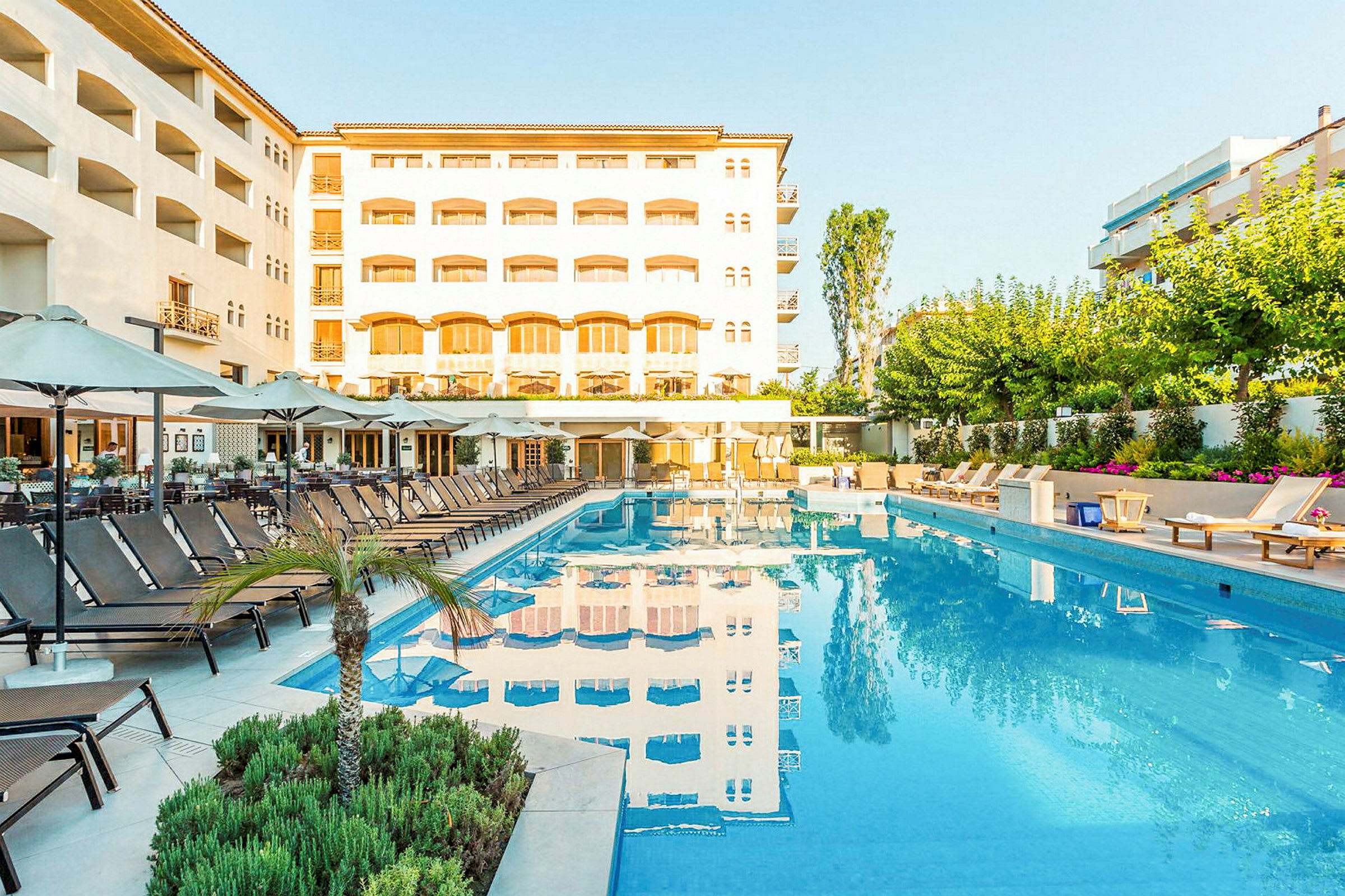 En udendørs swimmingpool med liggestole og parasoller omkring foran et hotel. (AI-genereret tekst)