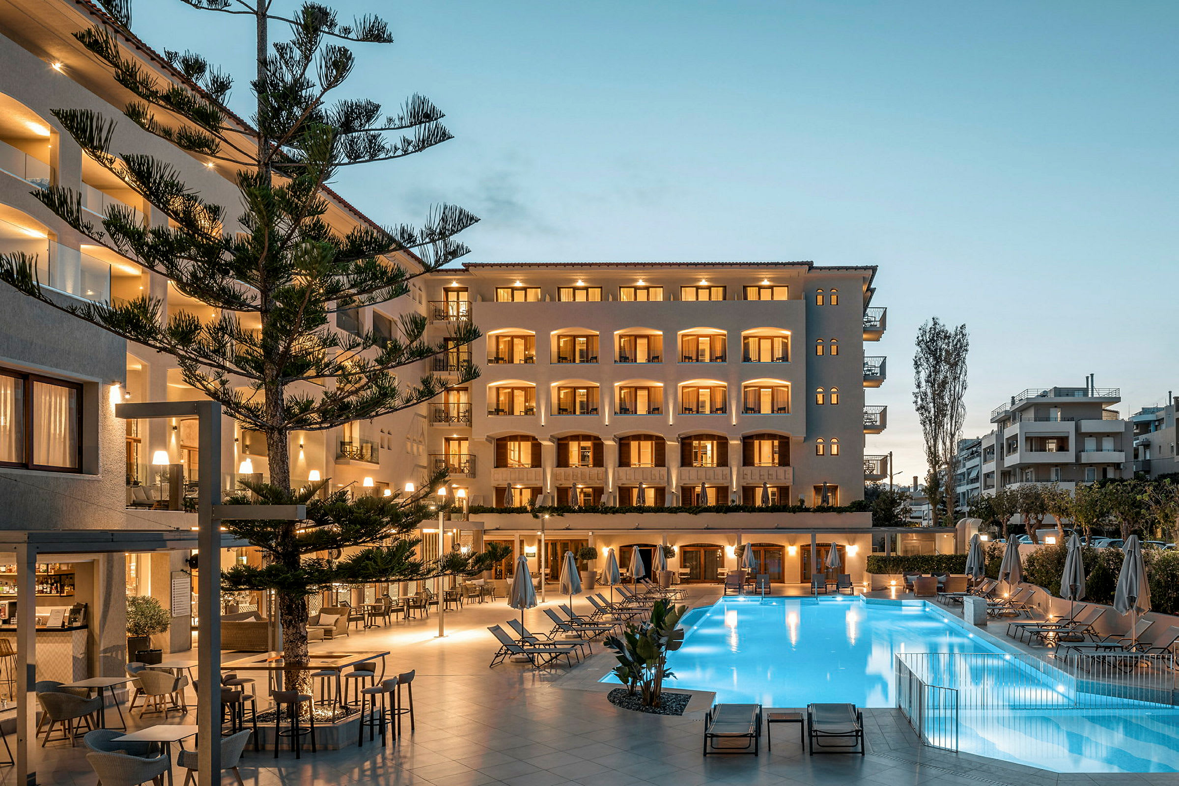 Udsigt over en oplyst hotelfacade med altaner foran en pool og udendørsområde om aftenen. (AI-genereret tekst)