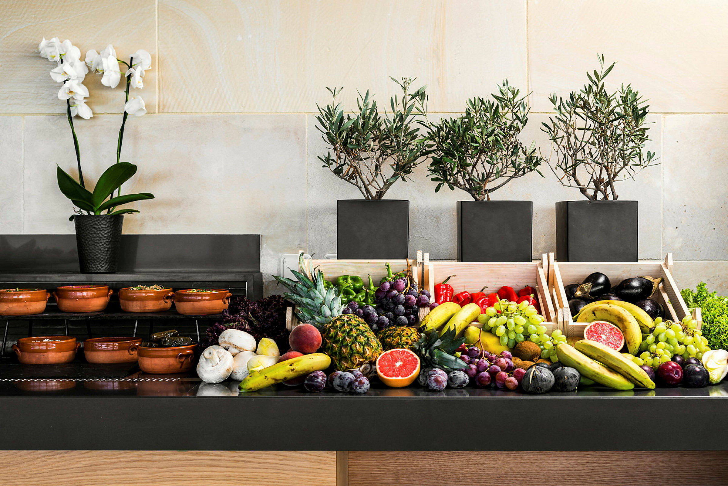 Buffetbord med frisk frugt og grøntsager samt oliventræer og en orkidé. (AI-genereret tekst)