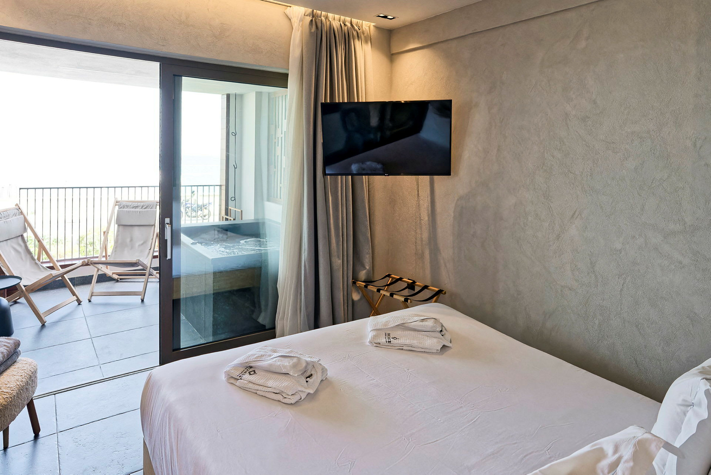 Moderne hotelværelse med hvid dobbeltseng, der fører ud til en balkon med liggestole og jacuzzi. (AI-genereret tekst)