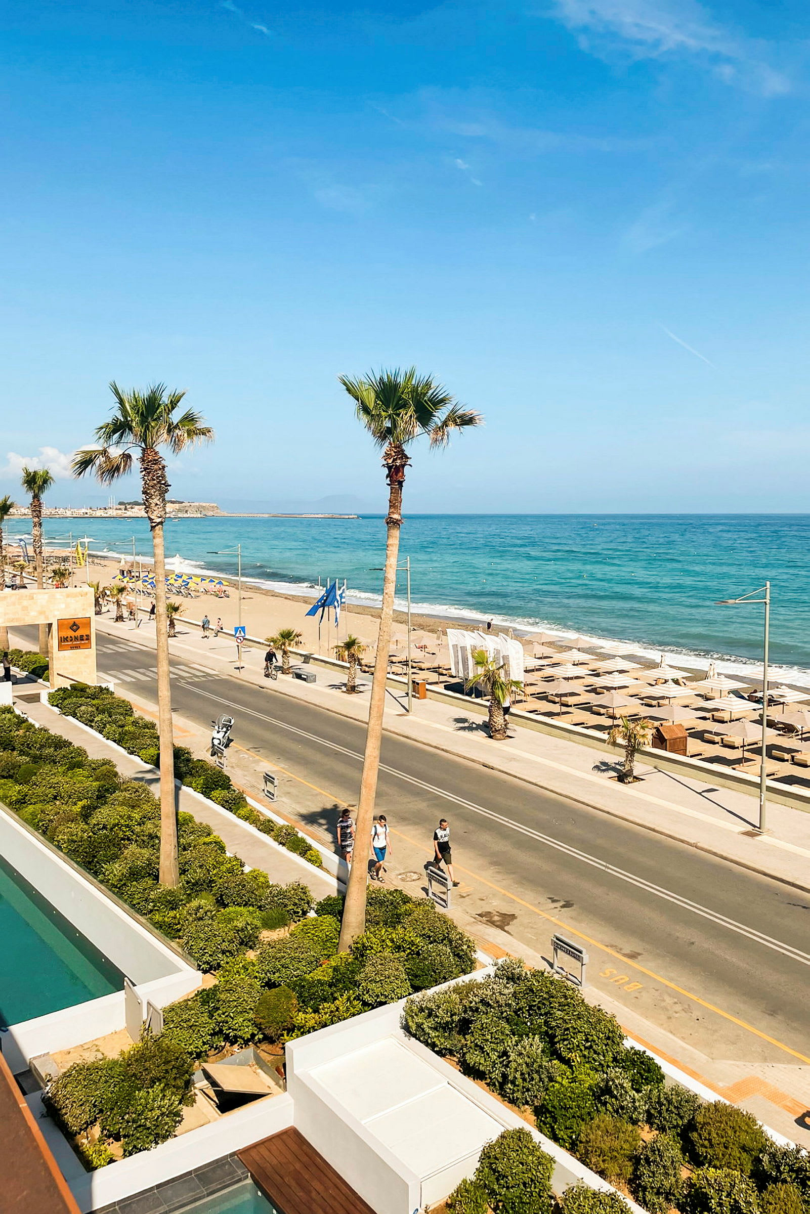 Udsigt over en strandpromenade med palmer, en vej og stranden ved havet. (AI-genereret tekst)