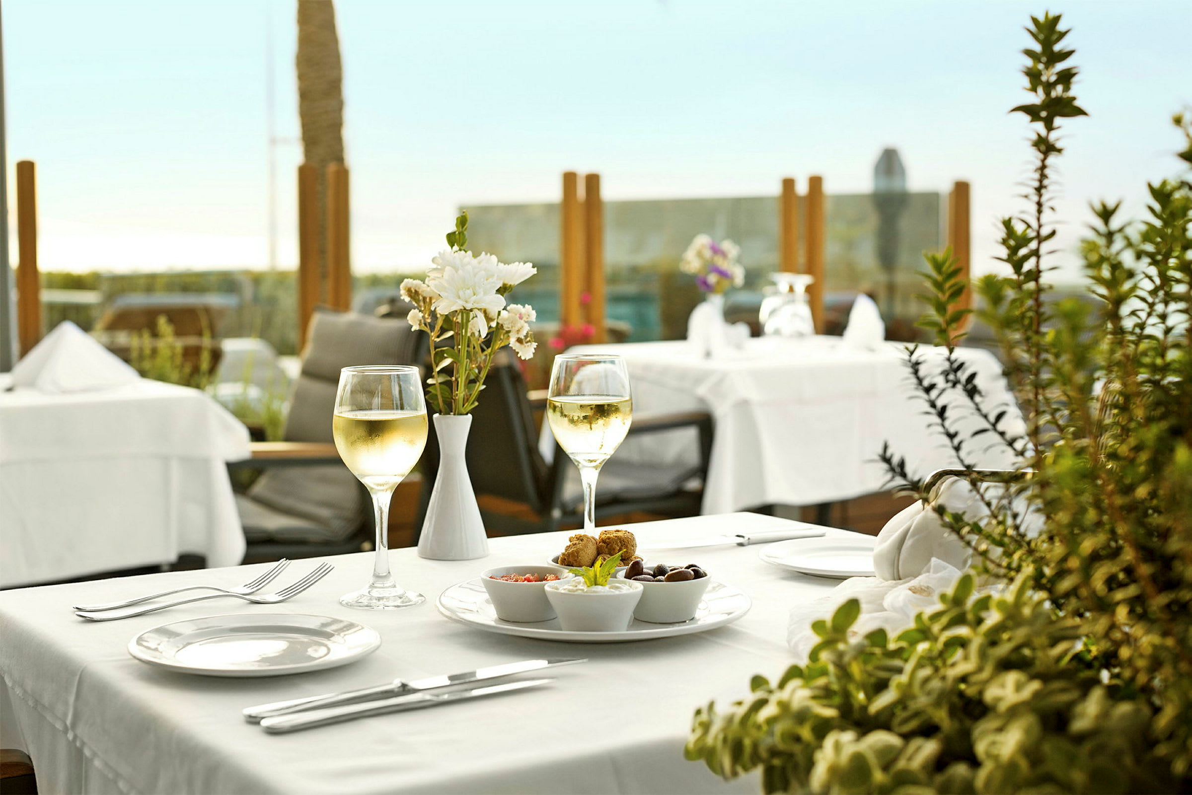 Et dækket restaurantbord med et glas hvidvin, blomsterdekoration og tallerken med snacks. (AI-genereret tekst)