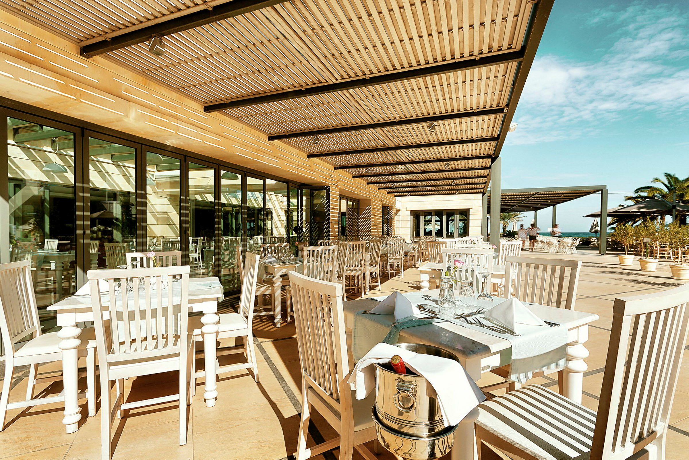 Udendørs restaurant med hvide borde og stole samt pergola der skygger. (AI-genereret tekst)