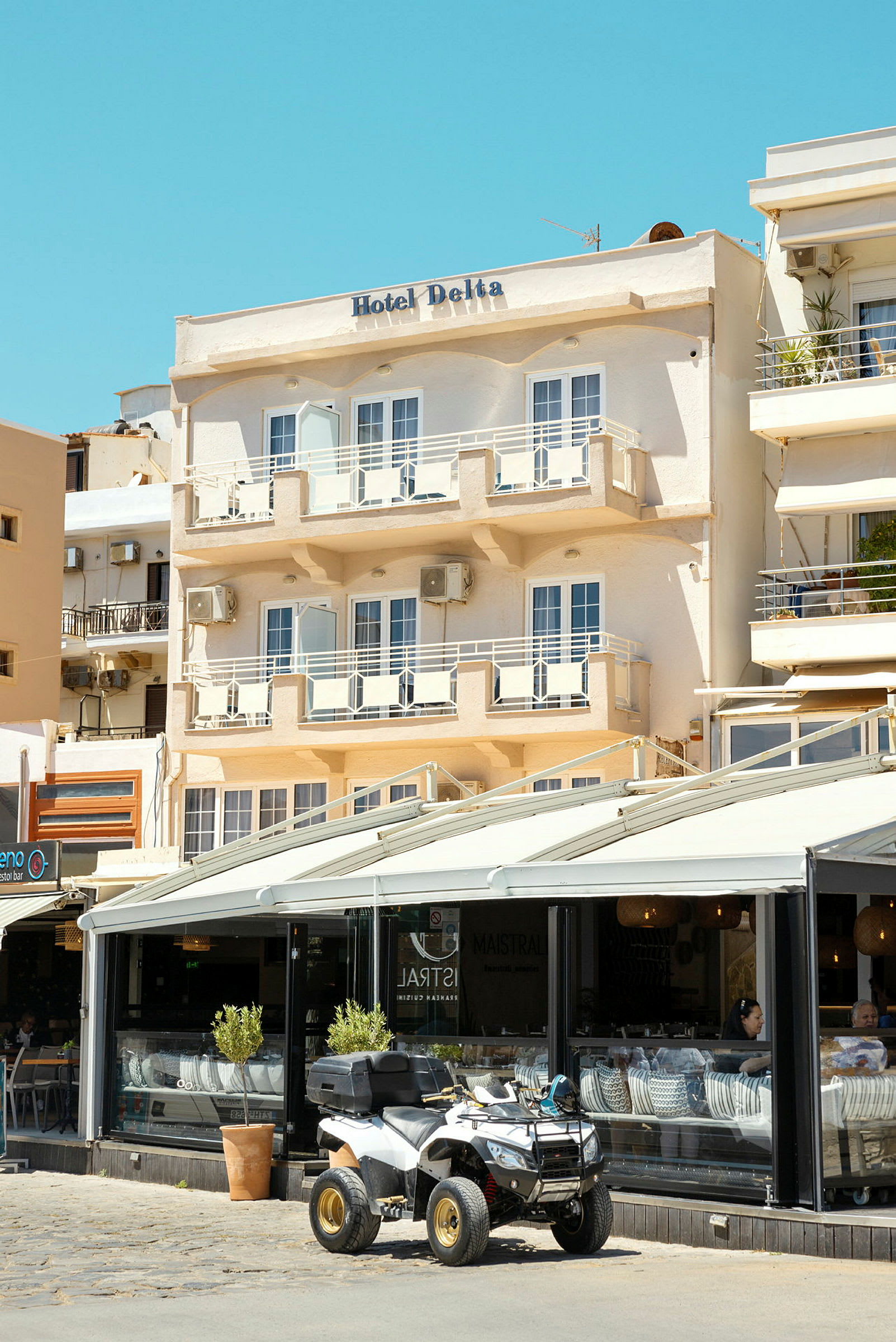 Facaden af et hotel ved navn "Hotel Delta" med altaner og en café i stueetagen, hvor en firehjulet motorcykel er parkeret udenfor. (AI-genereret tekst)