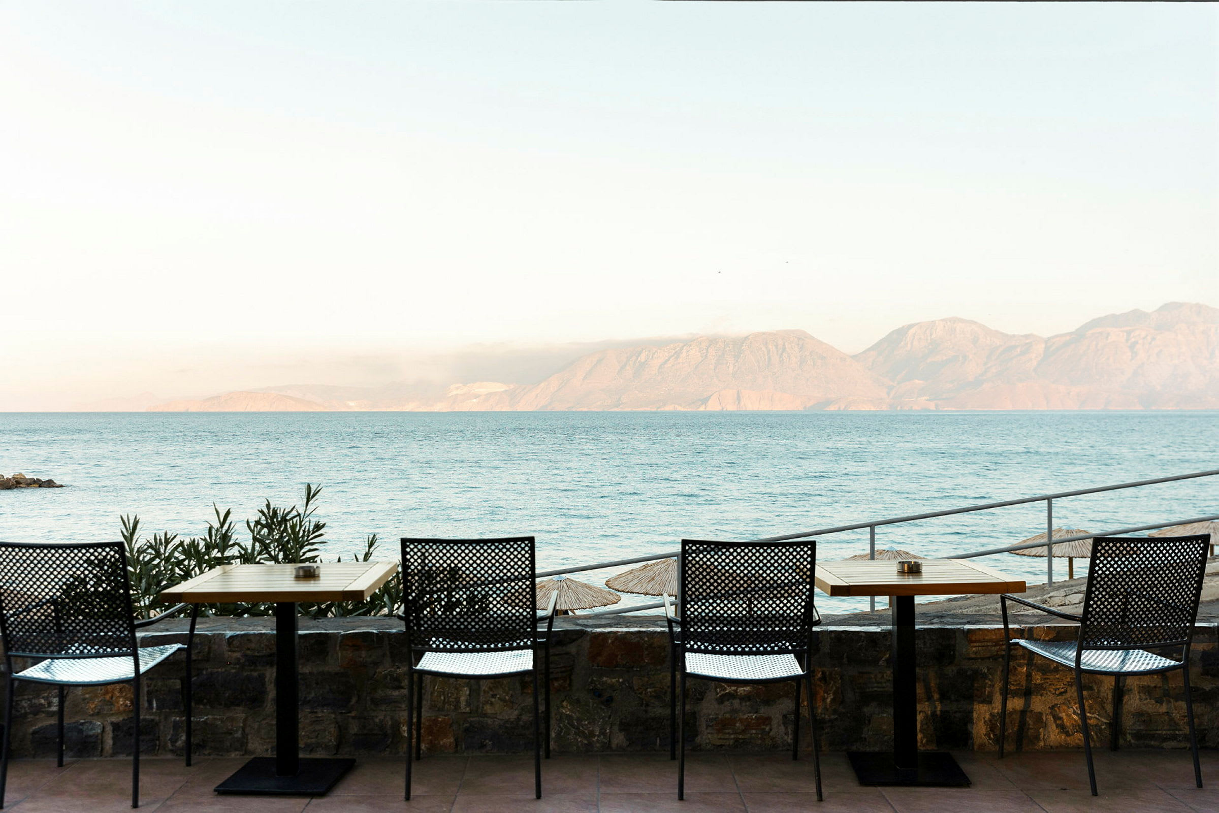 Billede af en udendørs restaurant med borde og stole samt udsigt til havet med bjerge i baggrunden. (AI-genereret tekst)