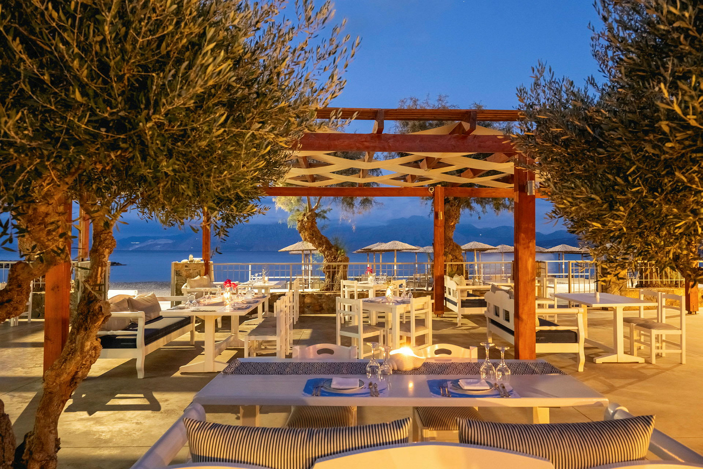 En udendørsrestaurant med siddepladser under en træpergola og udsigt over havet. (AI-genereret tekst)