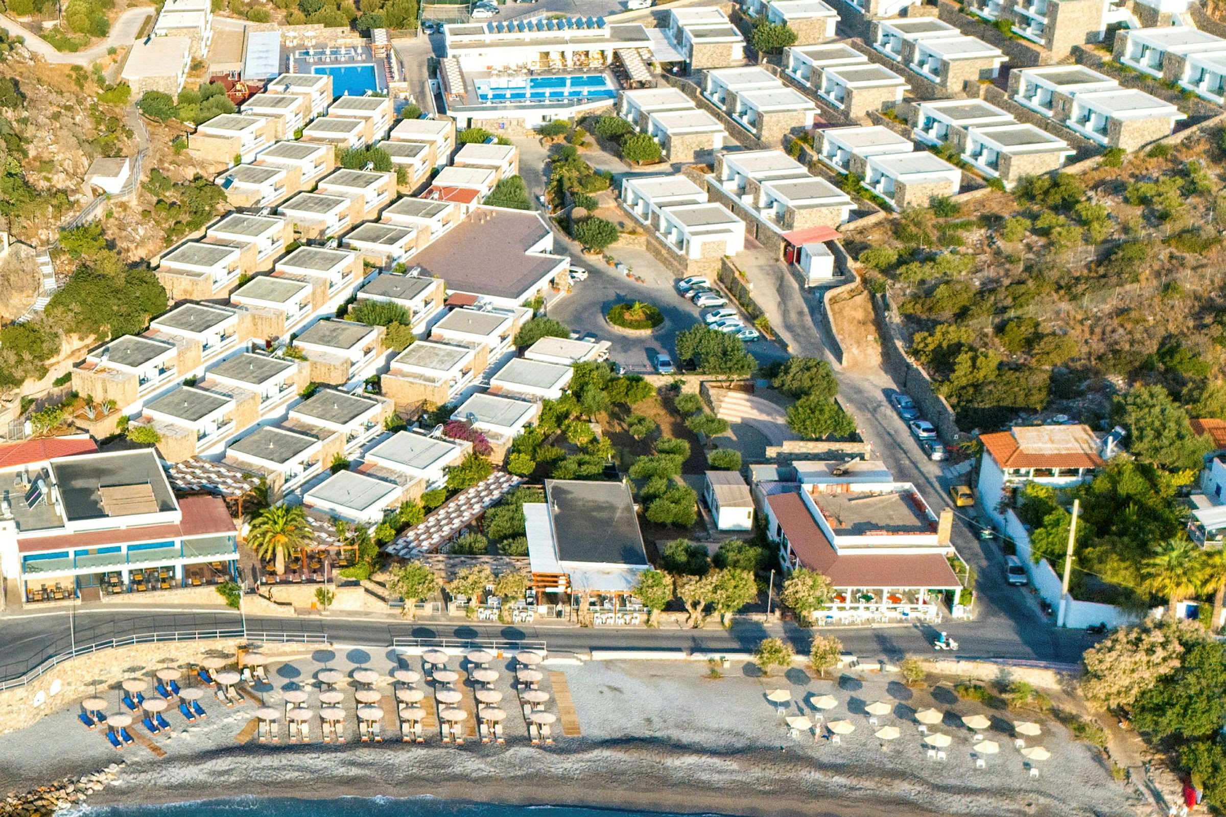 Luftfoto af et feriested med bygninger, pools og en strand med parasoller. (AI-genereret tekst)