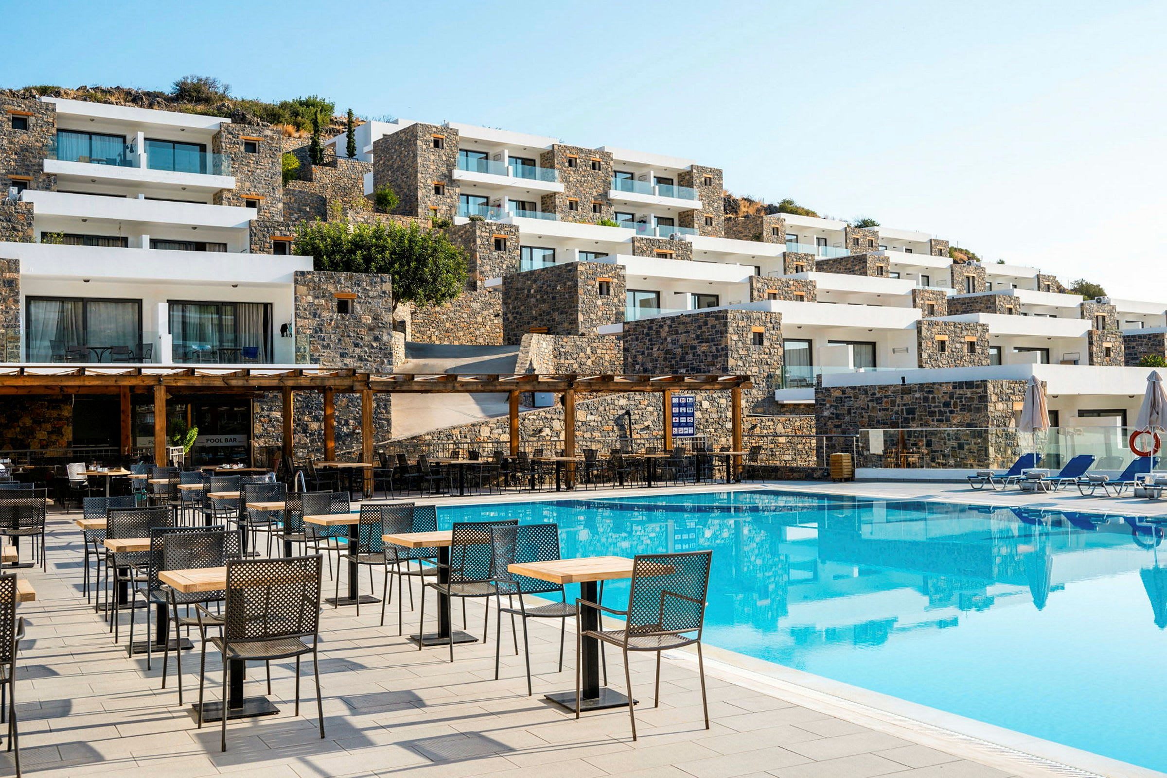 Billede af hotellets udendørsområde med pool, borde og stole samt stenbygninger i baggrunden. (AI-genereret tekst)