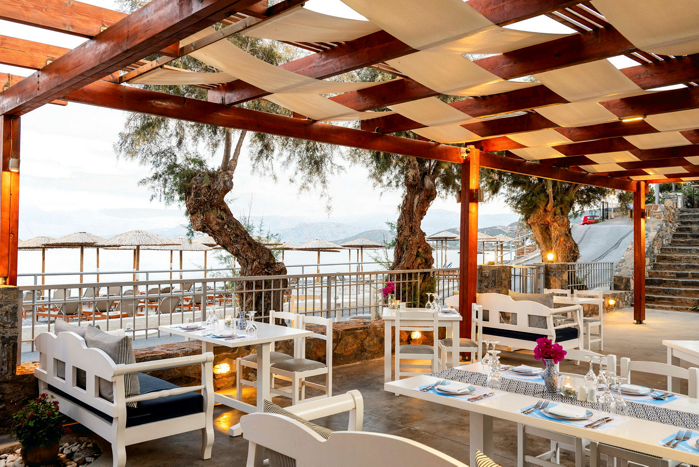 Restaurant med hvide borde og stole under en træpergola med udsigt over havet. (AI-genereret tekst)