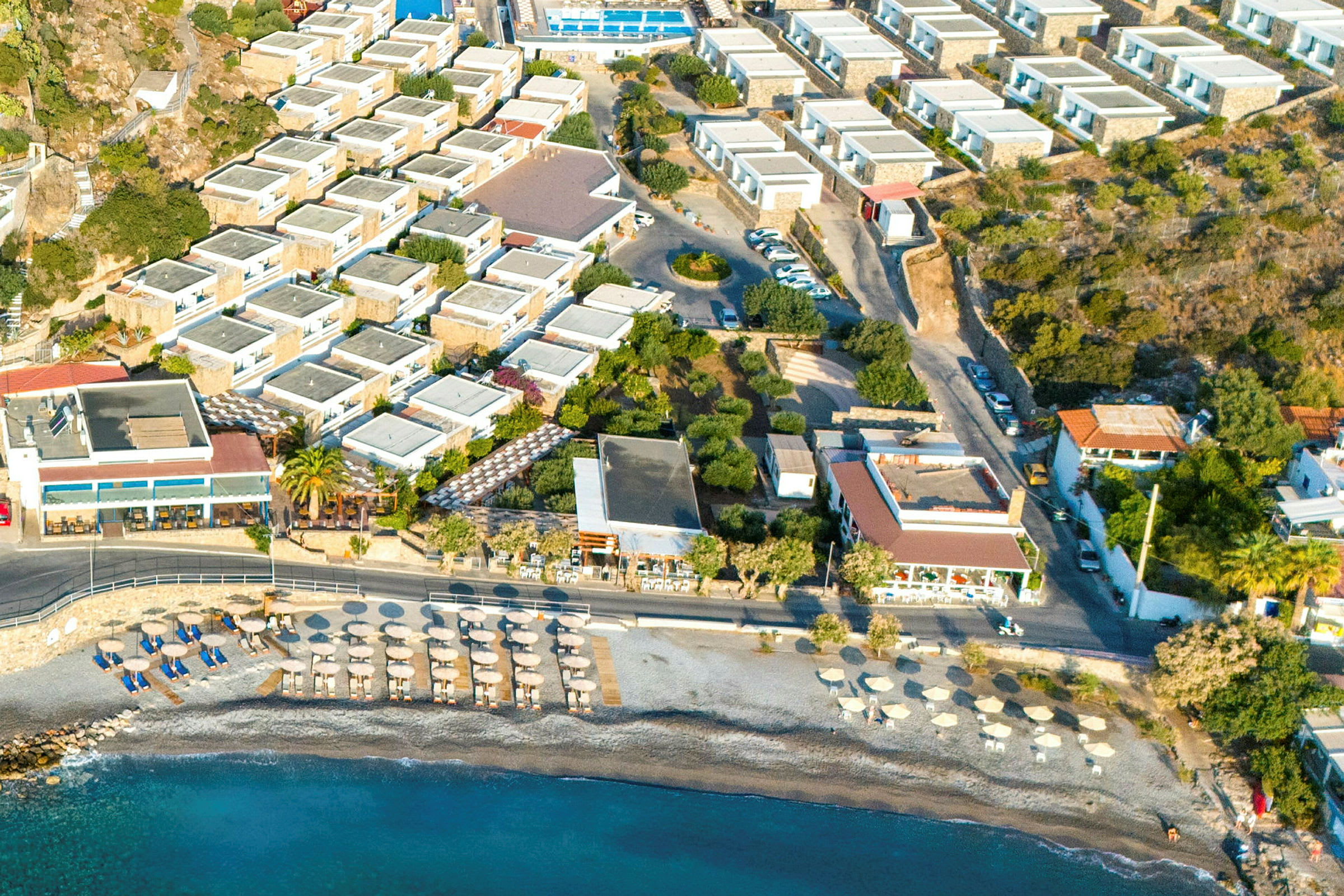 Luftfoto af hotelområde med swimmingpool, parkeringsplads og nærliggende strand med parasoller. (AI-genereret tekst)