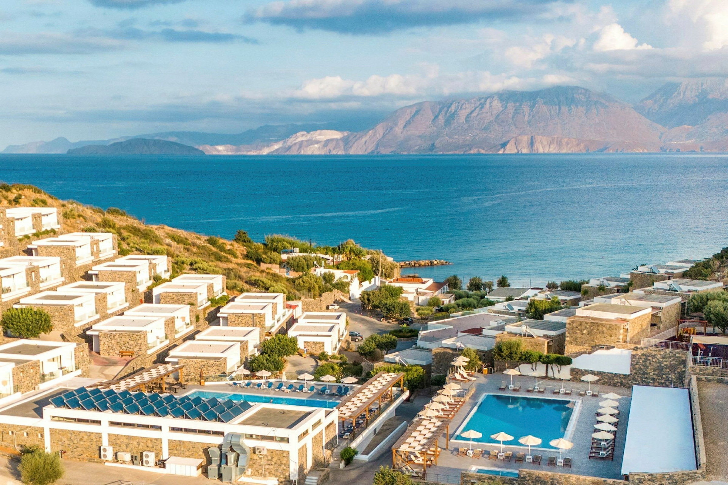 Billede af et kystnært resort med hotelbygninger på skrånende terræn, omgivet af grønne områder og bassiner, med hav og bjerge i baggrunden. (AI-genereret tekst)