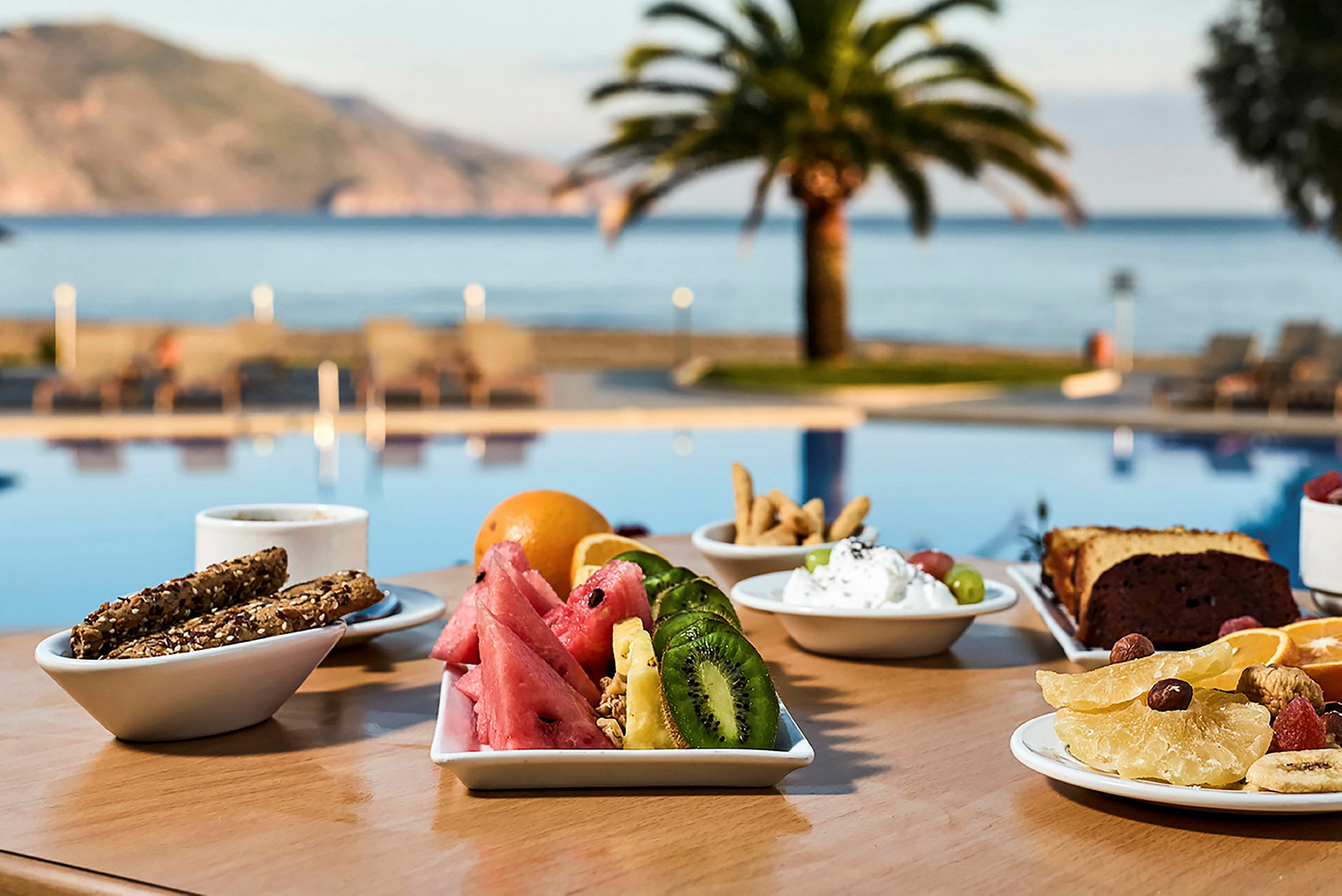 En morgenmad serveres på et bord tæt på en pool med udsigt over havet og palmer. (AI-genereret tekst)