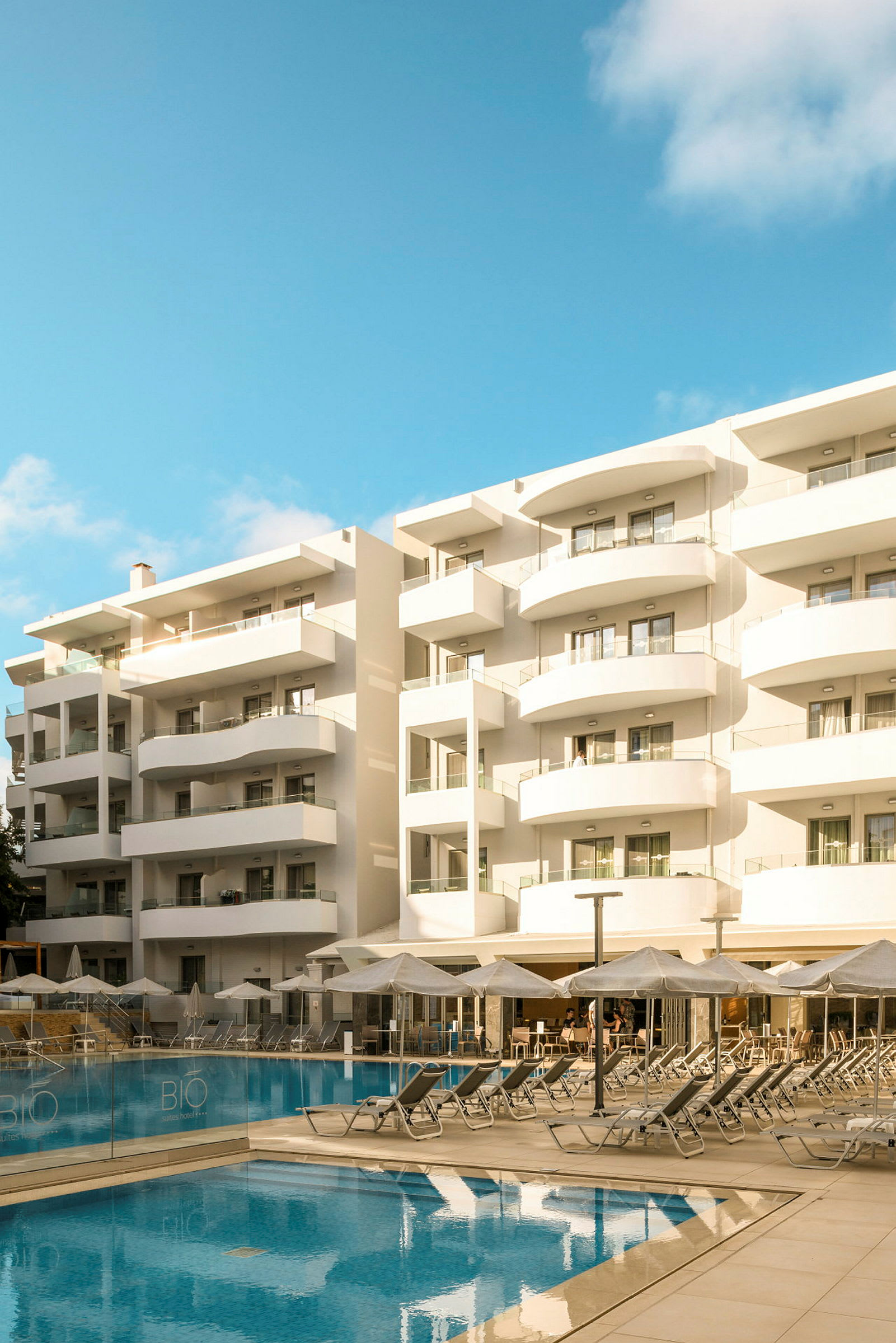 Et hotel med hvide altaner ved en pool med liggestole og parasoller. (AI-genereret tekst)