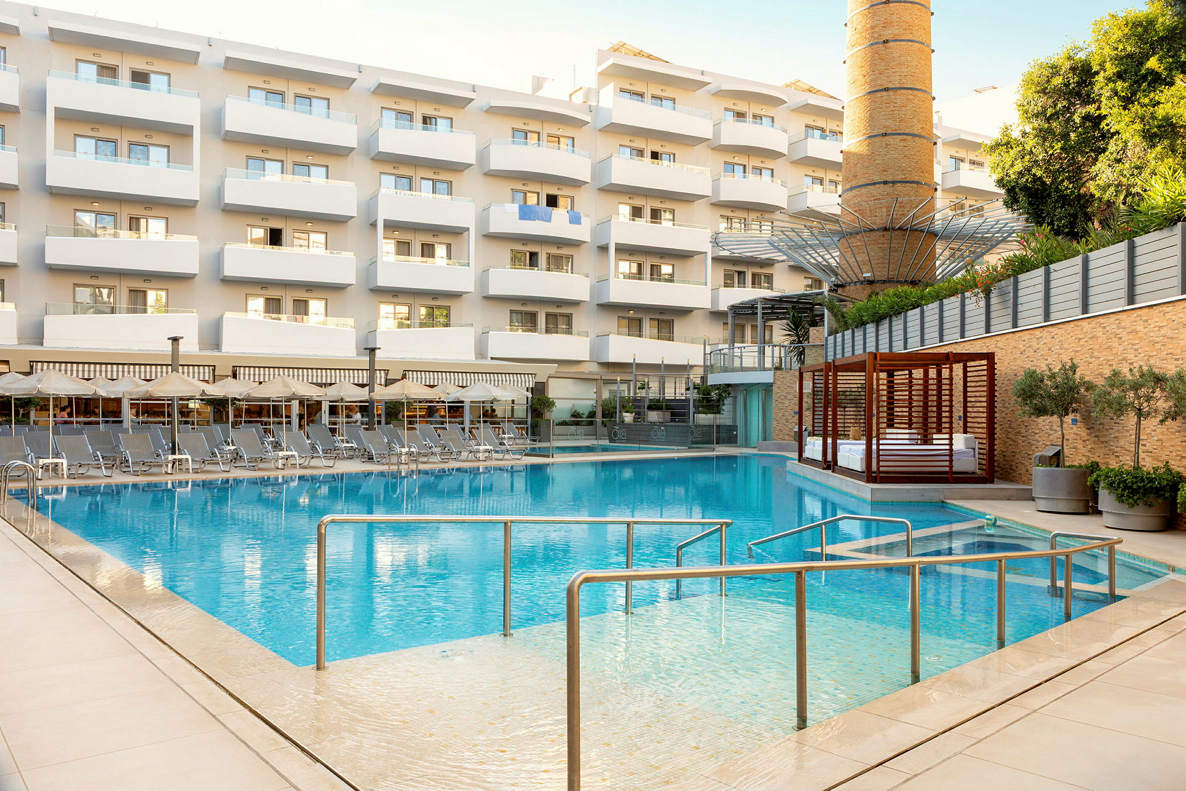 En udendørs pool foran en flerboligbygning med altaner. Poolområdet er omgivet af liggestole og parasoller. (AI-genereret tekst)