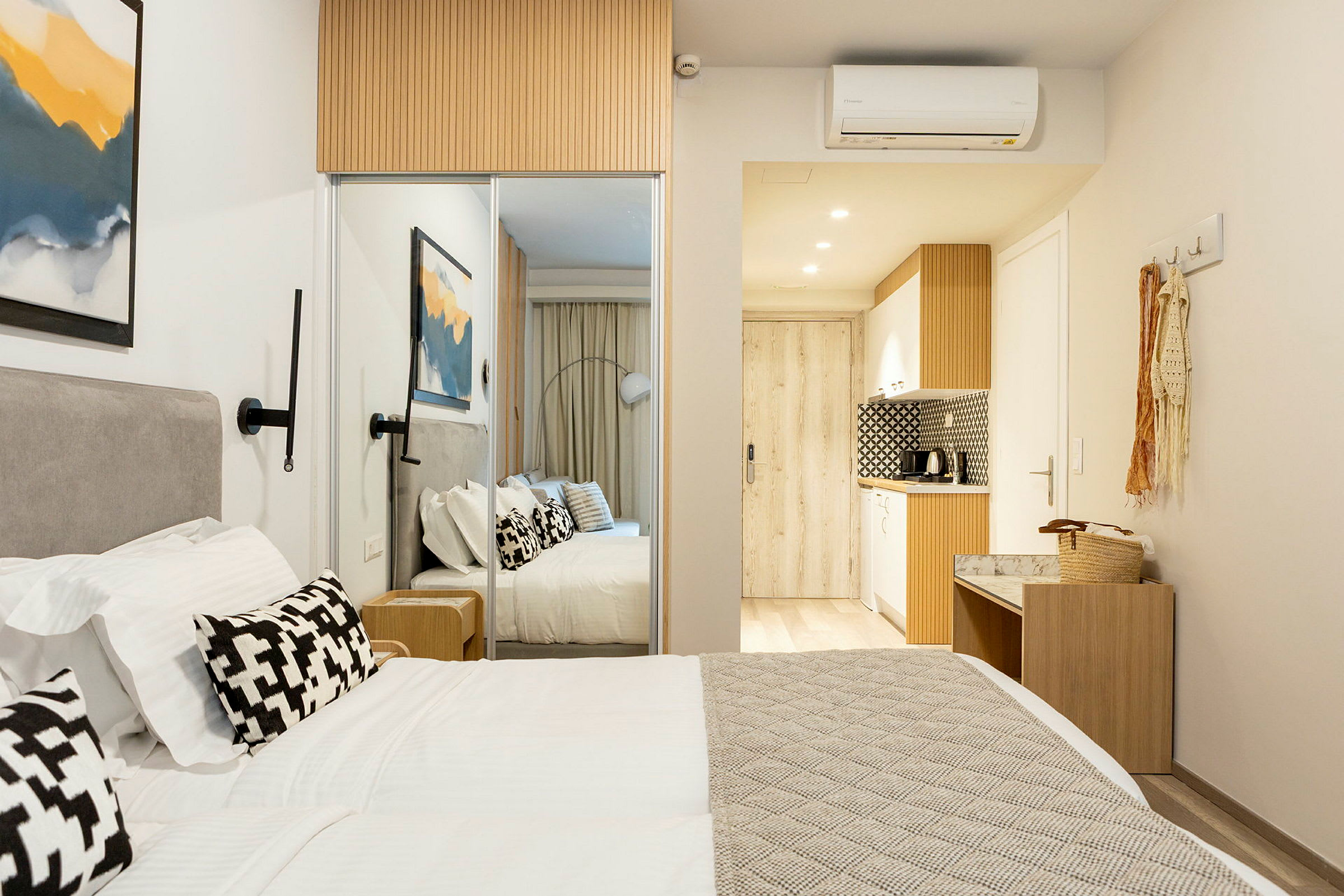 Et moderne hotelværelse med en dobbeltseng, hvide sengelagner og dekorative puder. På væggen hænger et abstrakt maleri, og i baggrunden ses et lille køkken. (AI-genereret tekst)