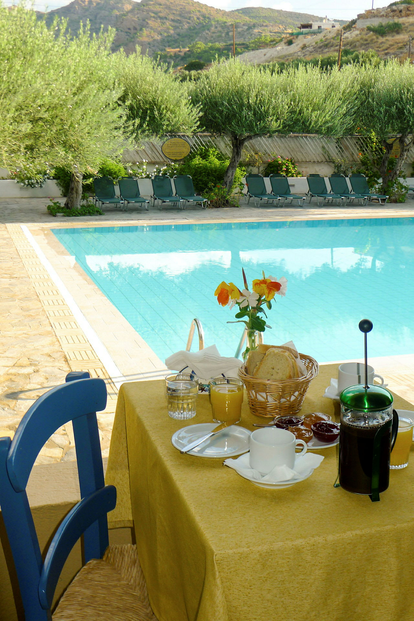 Et morgenbord med udsigt til en pool og liggestole, samt oliventræer og bjerge i baggrunden. (AI-genereret tekst)