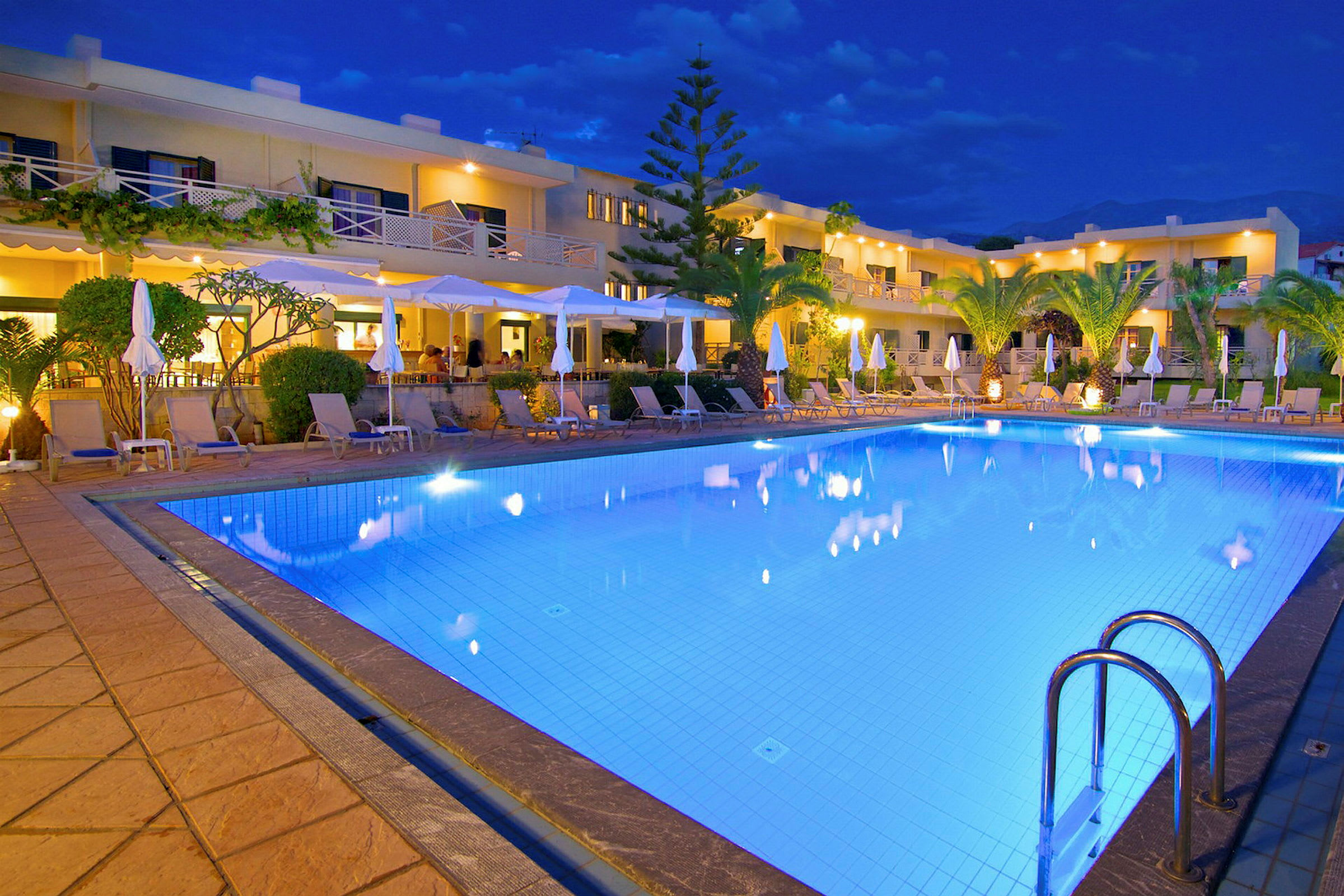 Aftenbillede af et oplyst poolområde foran et hotel med liggestole omkring poolen. (AI-genereret tekst)