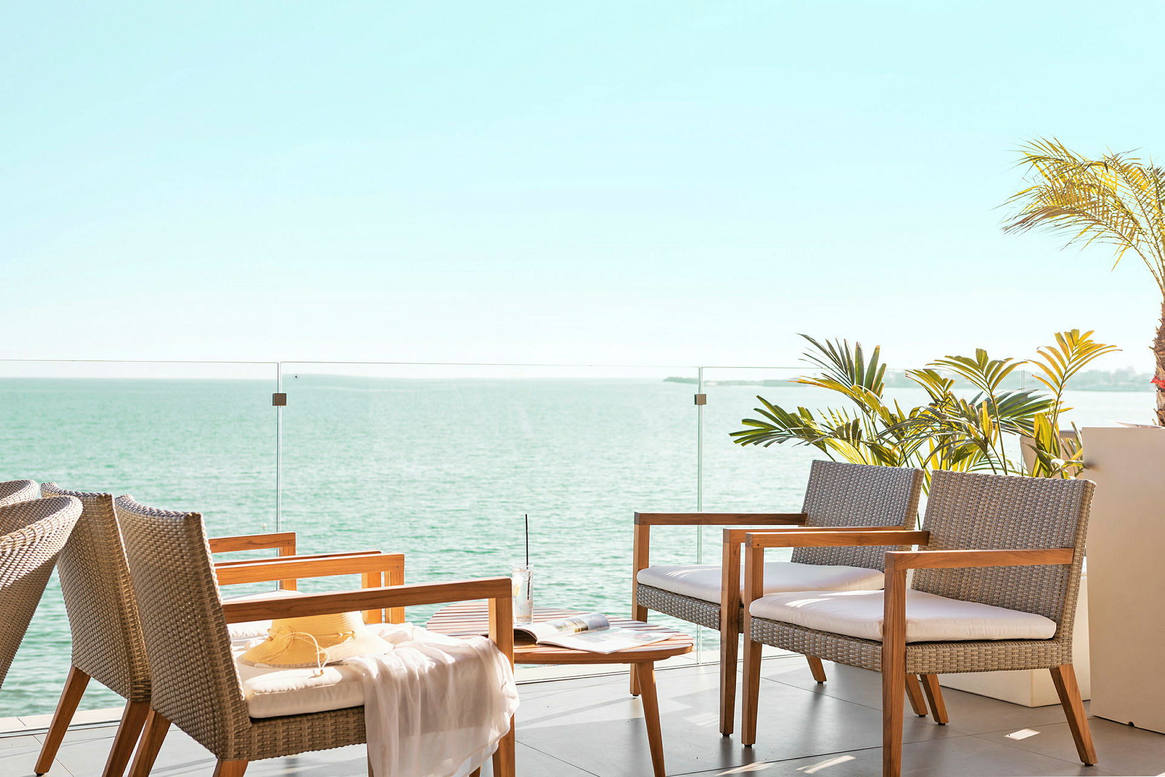 En siddegruppe med stole og bord ved en terrasse med glasrækværk. I baggrunden ses havet og himlen. (AI-genereret tekst)